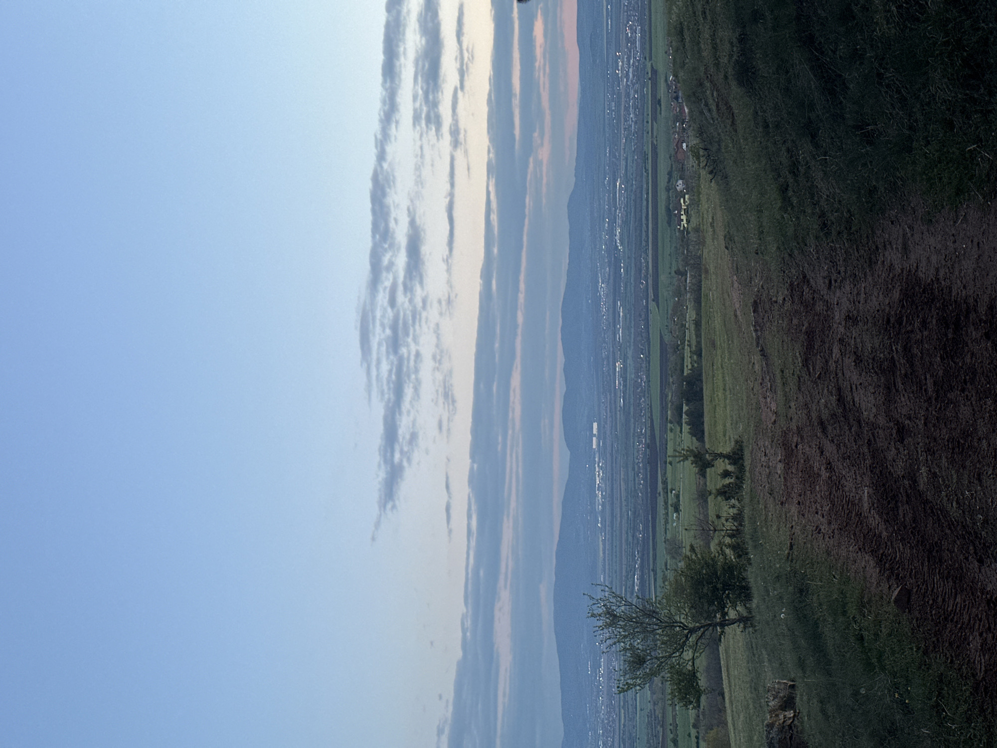 The scene presents a landscape viewed from a high vantage point in Balsha, Bulgaria, near the vicinity of the Balkan Mountains.
**Sky:** The left portion of the image shows a light blue sky, transitioning to streaks of grey, white, and pink clouds.
**Mountains and City:** A distant range of mountains forms a barrier above the city. The cityscape below is a mixture of residential and commercial buildings.
**Vegetation:** The right side of the image depicts grassy fields and trees. A brown dirt pathway runs through the field.