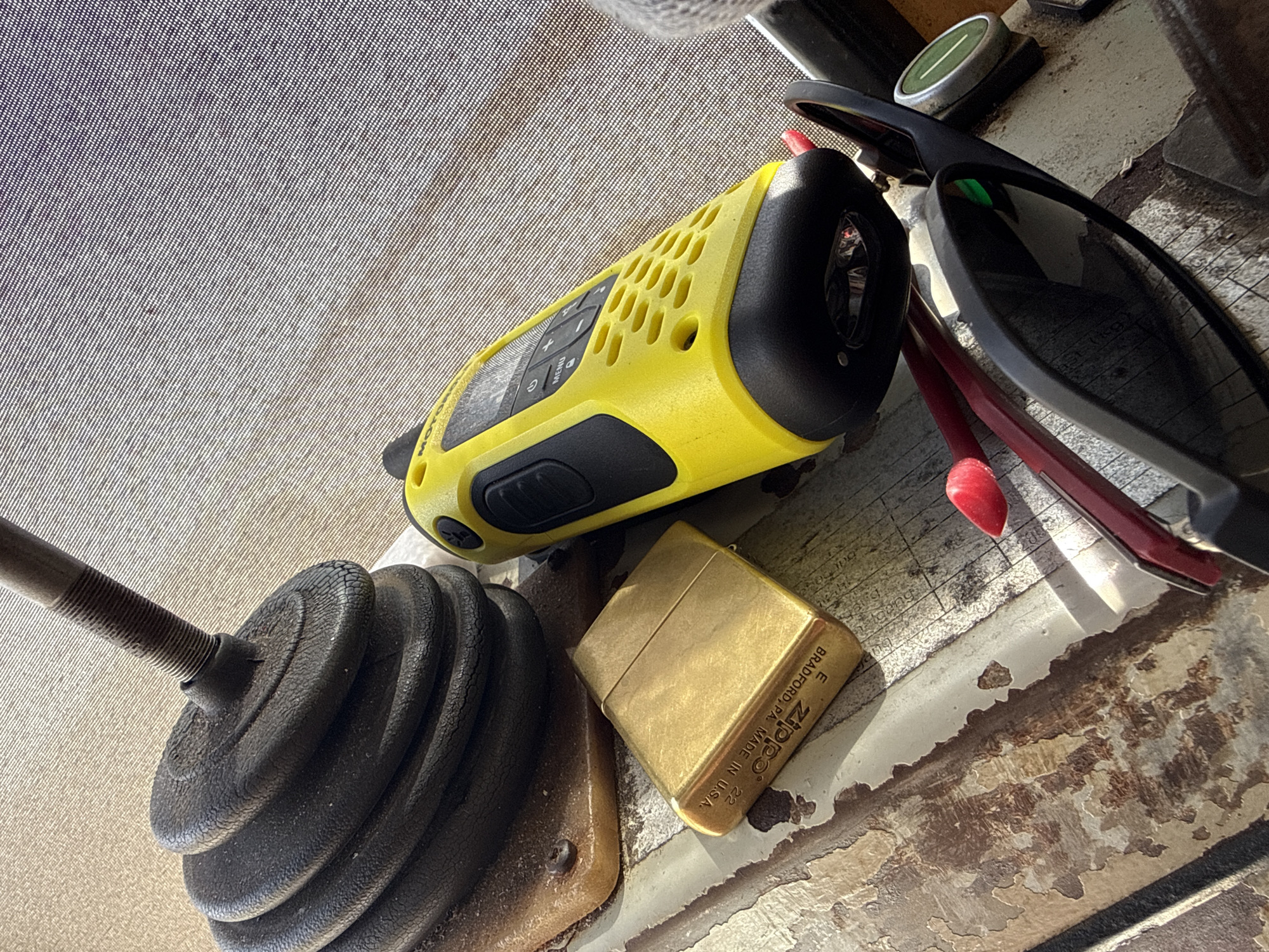 The image displays a collection of objects placed on a textured surface, possibly a table or a boat deck, given the Mikołajki, Poland location context. The dominant subjects are a bright yellow and black walkie-talkie, a brass Zippo lighter, a black weight plate with a threaded metal rod attached, and a pair of dark-rimmed sunglasses with a red accent visible. The walkie-talkie has a grid of ventilation holes and buttons, with the brand name "Motorola" partially visible. The Zippo lighter has text inscribed on it, including "BRADFORD, PA. MADE IN U.S.A.", and "ZIPPO". The weight plate appears to be made of black rubber or plastic and is stacked in several layers.
The lighting suggests it's daytime, with sunlight casting shadows and highlighting the textures of the objects and the surface. The surface itself has a weathered or distressed appearance, with peeling paint or material in some areas, revealing brown undertones. The scene is static, with no activity depicted. The overall impression is one of personal items laid out, perhaps before or after an outdoor activity. The objects are arranged closely together, creating a still-life composition.