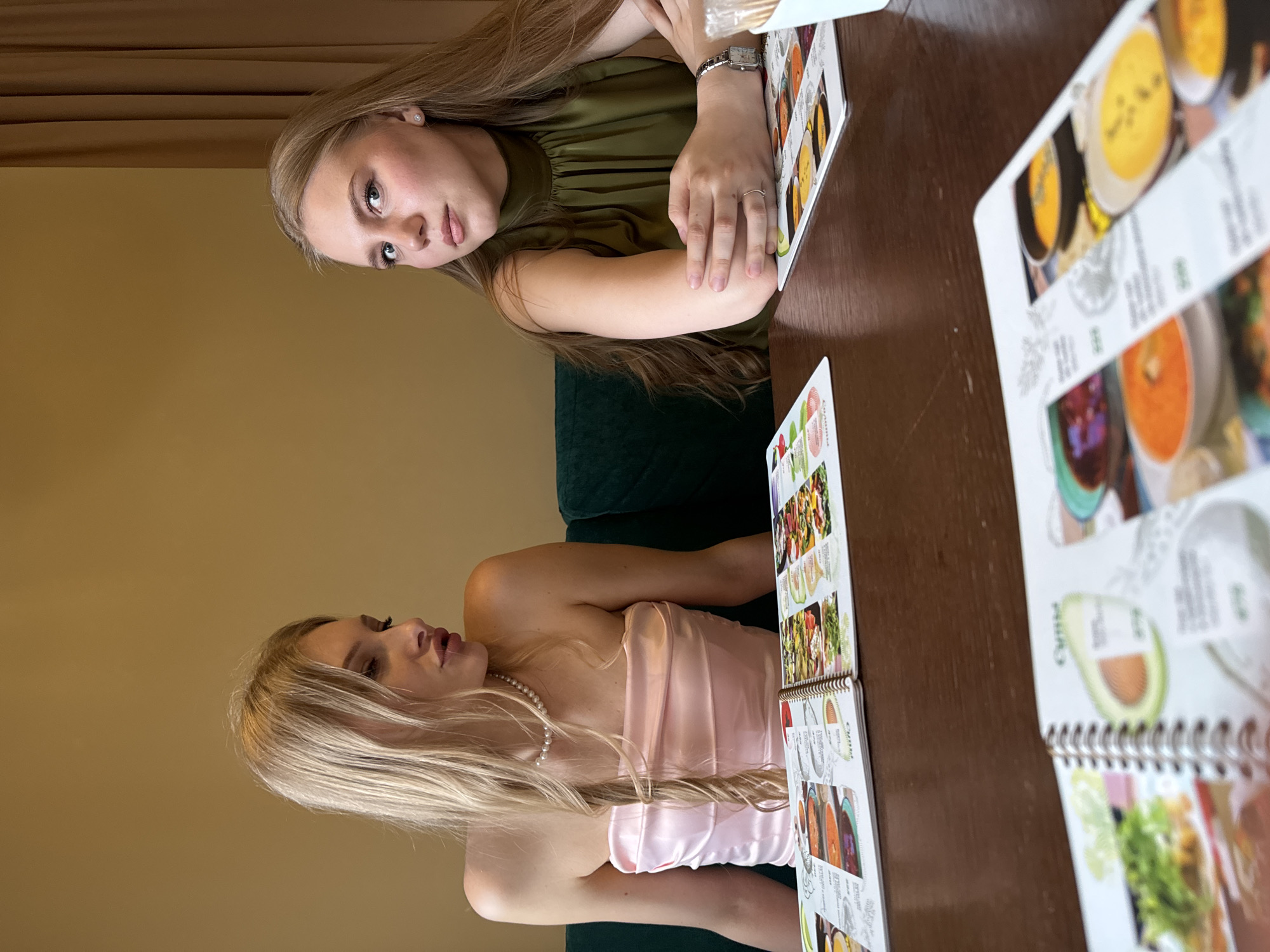 Two young women are seated at a wooden table, each examining a spiral-bound menu. The woman on the left, with blonde hair and wearing a pink strapless dress and a pearl necklace, is looking to the right. The woman on the right, with brown hair and wearing a green top, is looking towards the camera. The setting appears to be a restaurant or cafe with an upholstered green chair. The location is likely Perm, Russia, based on the available information.