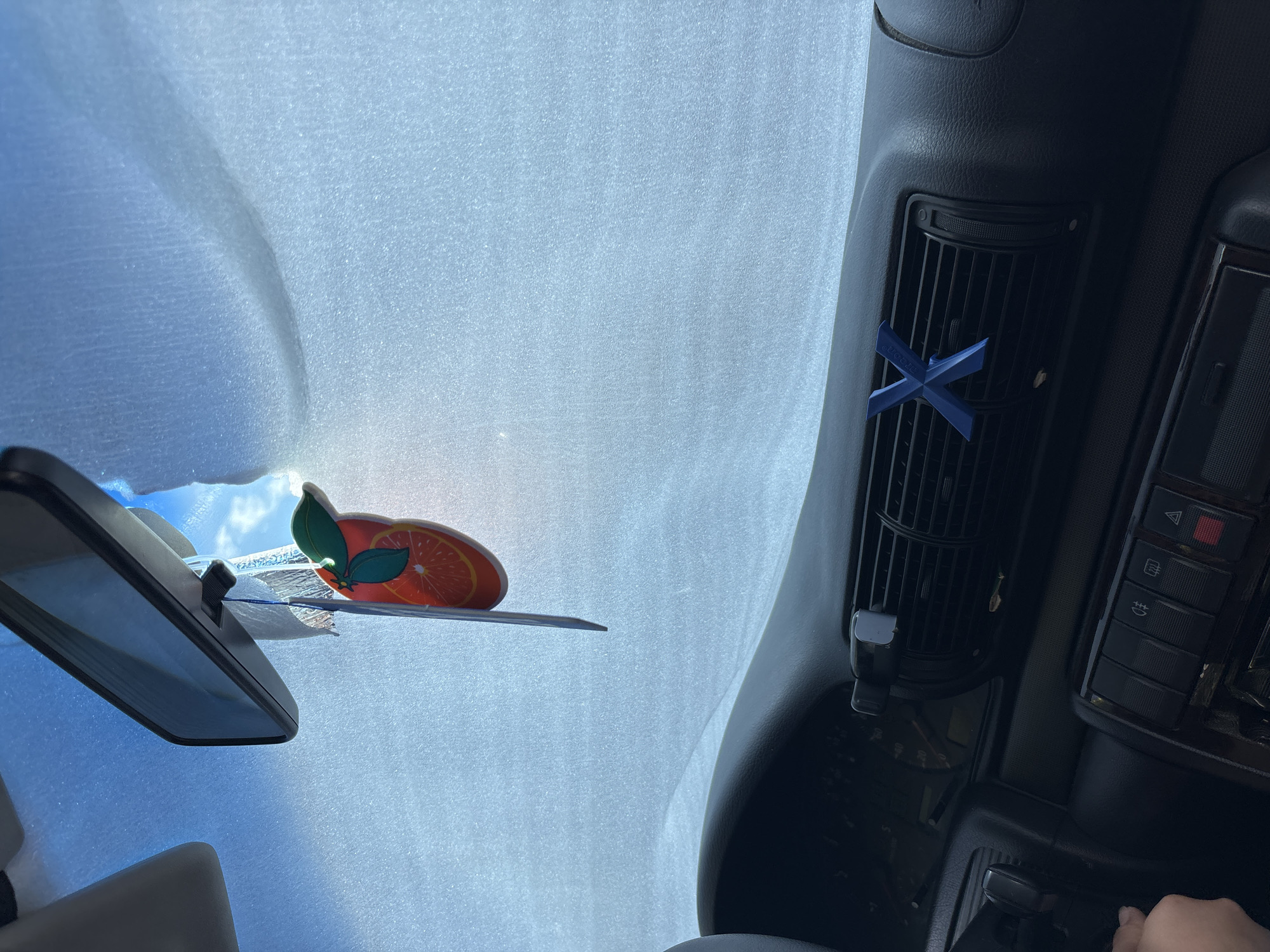 The image shows the interior of a car, likely in Varna, Bulgaria, looking forward through the windshield. A sunshade covers the majority of the windshield, with a tear in the top right corner revealing a bright blue sky. A rearview mirror is visible above the sunshade, with a dangling air freshener shaped like an orange slice hanging from it. On the dashboard, a blue "X" shaped object is clipped onto an air vent. The dashboard features various controls and buttons, including a hazard light button and other warning indicators. A hand is visible on the left side of the frame, suggesting a person is in the driver's seat. The scene appears to be captured during daylight, with sunlight filtering through the windshield.