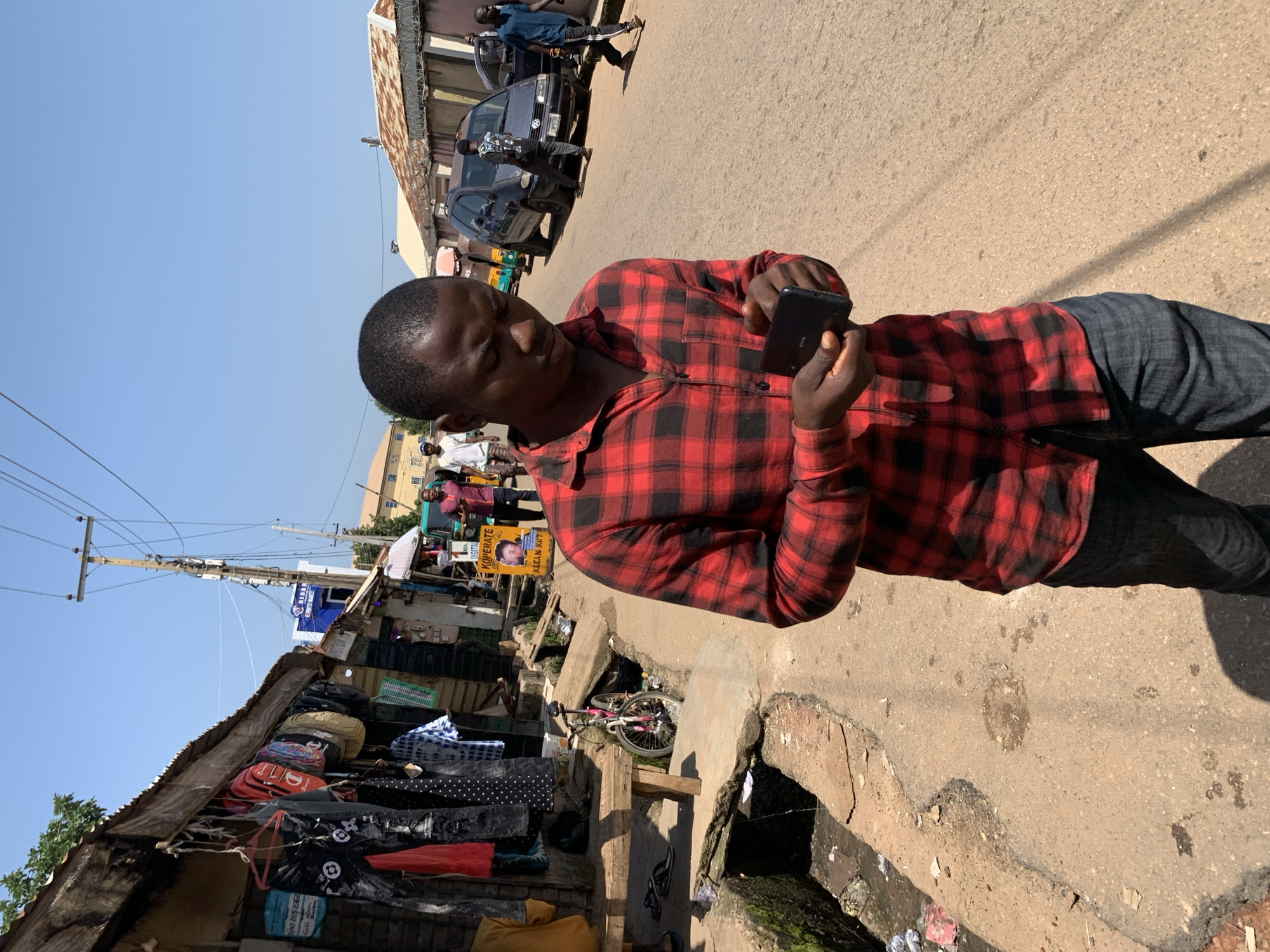 A young Black man wearing a red and black plaid shirt and dark blue jeans is walking down a dusty street, looking at a black smartphone in his hands. He is in the foreground, with a market stall to his left and a street with a few vehicles and other pedestrians to his right. The market stall is made of corrugated metal and wood and has various items of clothing and bags hanging from it. A bicycle is leaning against a concrete structure in front of the stall. The street is paved and has a drainage ditch on the left side. In the background, there are buildings, a blue sign with white lettering, and a utility pole with many wires. The sky is clear and blue. The scene appears to be in an urban environment in Jos, Nigeria, likely a marketplace area.