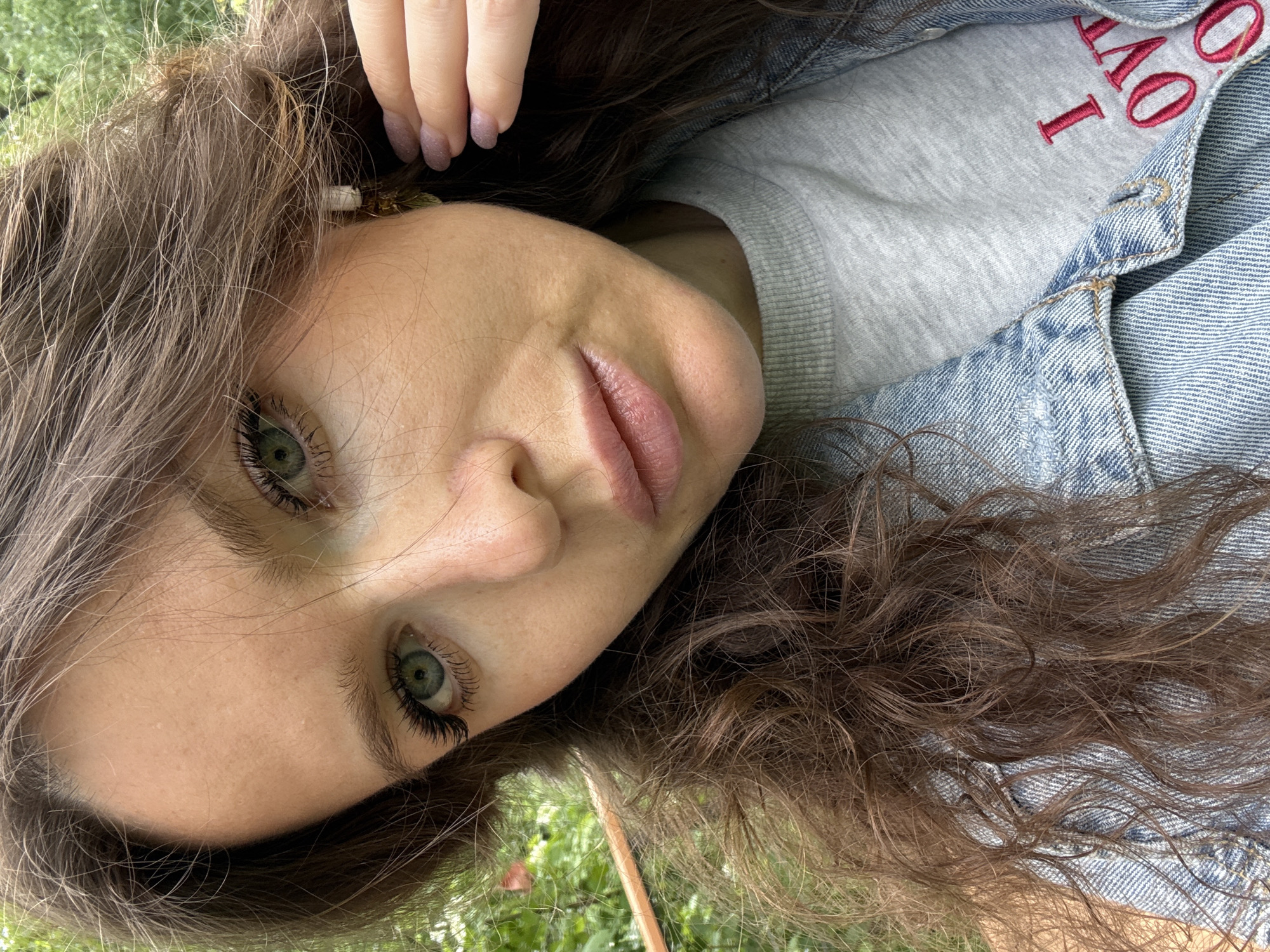 A woman with fair skin, green eyes, and brown hair is captured in a close-up shot, her face tilted to the side. She wears a light gray sweatshirt under a denim jacket, with the red embroidered word "LOVE" partially visible on the sweatshirt. Her fingernails are painted with a light-colored polish. The background suggests an outdoor setting with greenery, possibly grass, visible at the bottom of the frame. The location is not discernible, so it can't be confirmed in Moscow, Russia.
