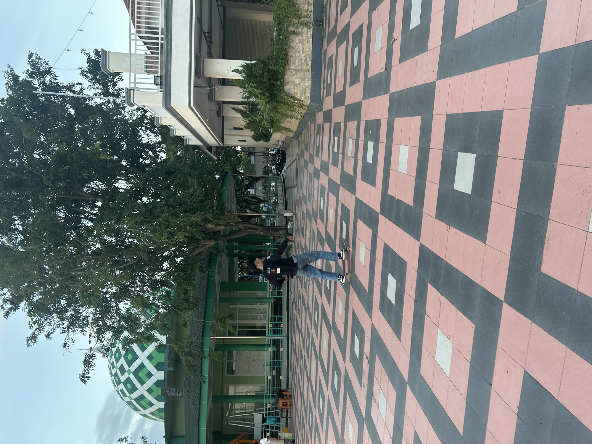 In Bantaeng, Indonesia, on a sunny day, a young man in a black long-sleeved shirt and blue jeans is skateboarding on a patterned courtyard. The courtyard features a repeating design of pink and dark grey tiles, with lighter grey squares interspersed. To the left, a green building with white window frames and a distinctive green dome with a white lattice pattern, likely a mosque, is visible. A large tree with dark green foliage stands between the courtyard and the mosque. To the right, a white building with columns and a railing on an upper level is partially visible. The sky is a pale blue with some clouds. In the background, several motorcycles are parked, and string lights are suspended overhead.
