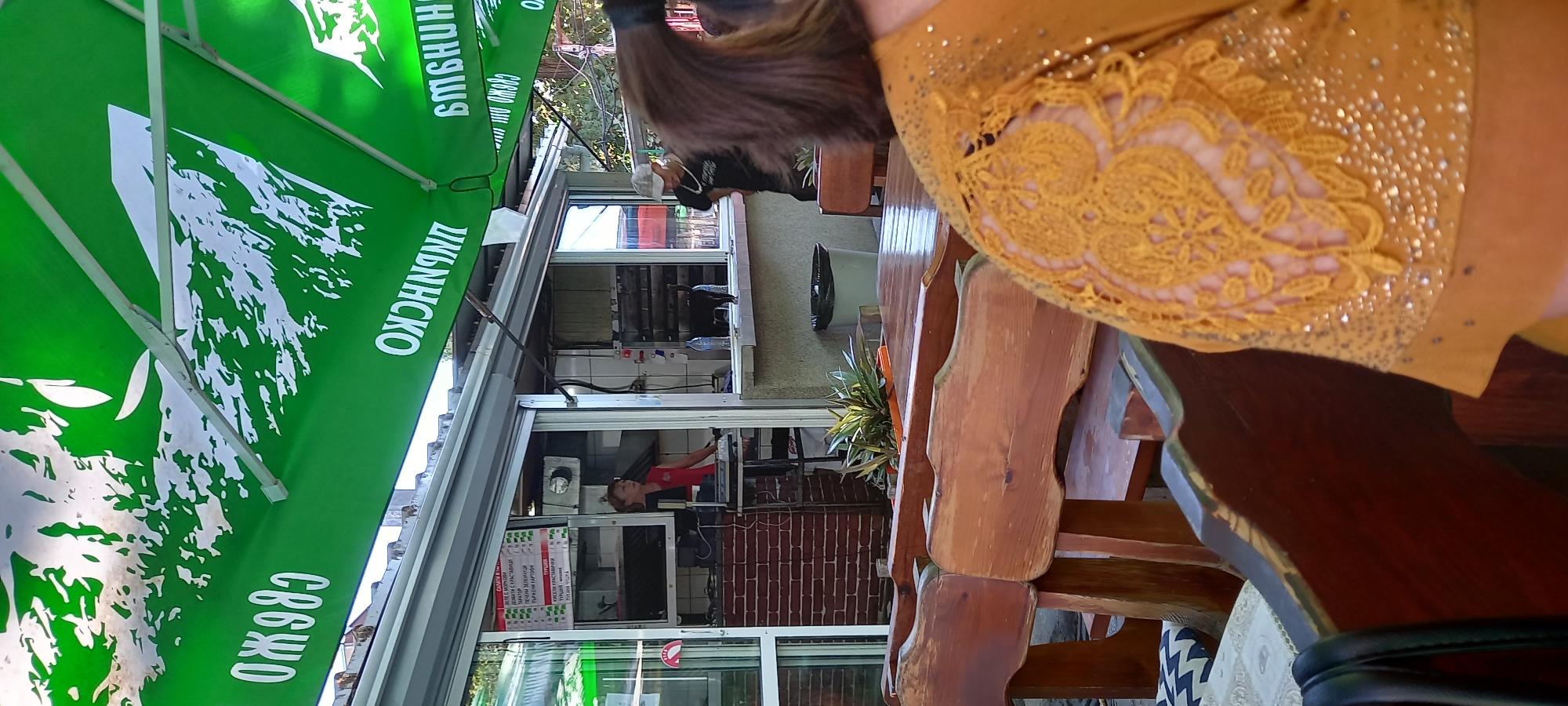 This image captures a view of an outdoor seating area at a fast-food establishment, likely located in Silistra, Bulgaria, as indicated by the Cyrillic lettering on the green umbrella that reads "ОЖДЯРО" and "ЛИПНИНСКО" and "БМБНЦН". In the background, a woman in a red and black shirt stands behind a counter, possibly taking an order. To her right, a black cat walks along the counter. The seating area features wooden tables and benches, with some patrons visible in the foreground, one wearing a distinctive yellow top adorned with intricate lace and embellishments. The overall scene suggests a casual dining experience on a bright day.