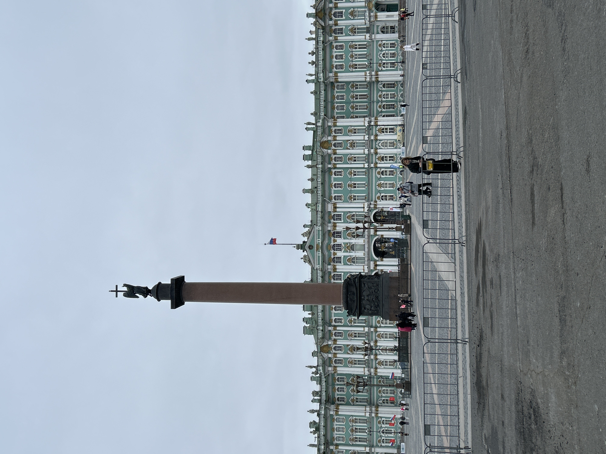 The image depicts a grand public square in Saint Petersburg, Russia, identified as Palace Square. The sky is overcast, suggesting a cloudy day with soft, even lighting.
The dominant architectural feature on the right is the magnificent Winter Palace, part of the State Hermitage Museum. It exhibits a distinctive Baroque facade painted in shades of light green/teal, accented with white columns and window frames, and intricate gold detailing. Numerous windows, both arched and rectangular, adorn its multi-story structure. A Russian flag (blue, white, red) flies prominently from a flagpole on the palace's roof, and smaller flags or banners are visible along its base.
In the center-left of the square stands the towering Alexander Column. This monolithic red granite column is crowned with a bronze angel holding a cross, a symbolic monument.
The expansive square is paved with a mix of dark asphalt and lighter stone sections. Dark metal barriers are arranged across the middle ground, likely used for crowd control or delineating event spaces.
Several individuals are scattered across the square, appearing to be tourists or visitors. They are casually dressed, with some wearing jackets, consistent with potentially cool, overcast weather. Some people are walking, while others are standing near the barriers or closer to the palace, observing the surroundings. One person in a dark jacket with a yellow item is visible near the barriers. No specific event is clearly taking place, suggesting general public access and sightseeing. There is no visible text in the image.