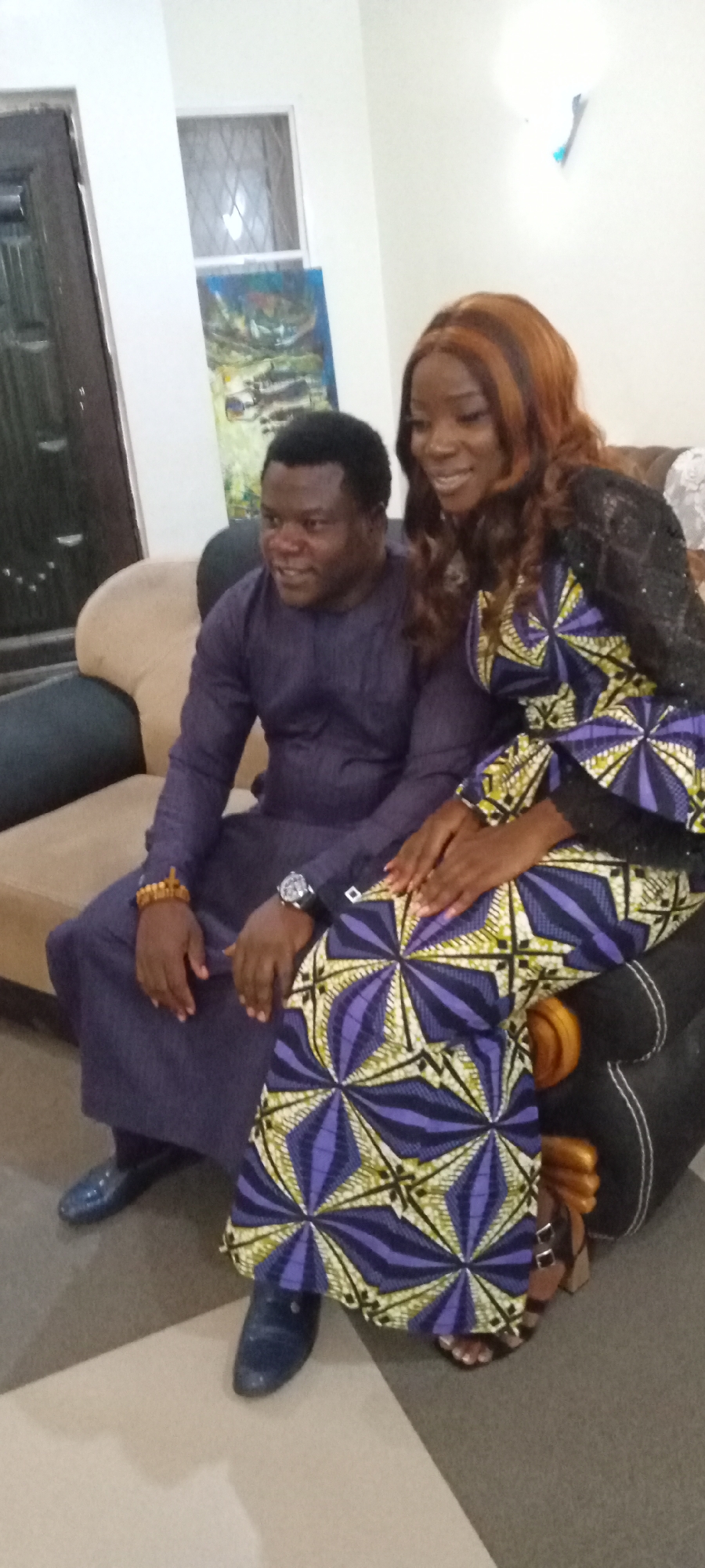 A medium shot shows a man and woman seated together on a couch. The man, positioned to the left, wears a long-sleeved purple tunic and matching shiny blue shoes. A gold bracelet adorns his left wrist, and a watch is visible on his left. The woman sits to his right, partially overlapping him. She is attired in a vibrant African print dress with purple, yellow, and white geometric patterns, complemented by a black lace top. Her hair is long and wavy, with streaks of light brown highlights. They are sitting on a brown couch. In the background, a painting hangs on the wall, and there is a dark-colored door. The location and city are unknown.