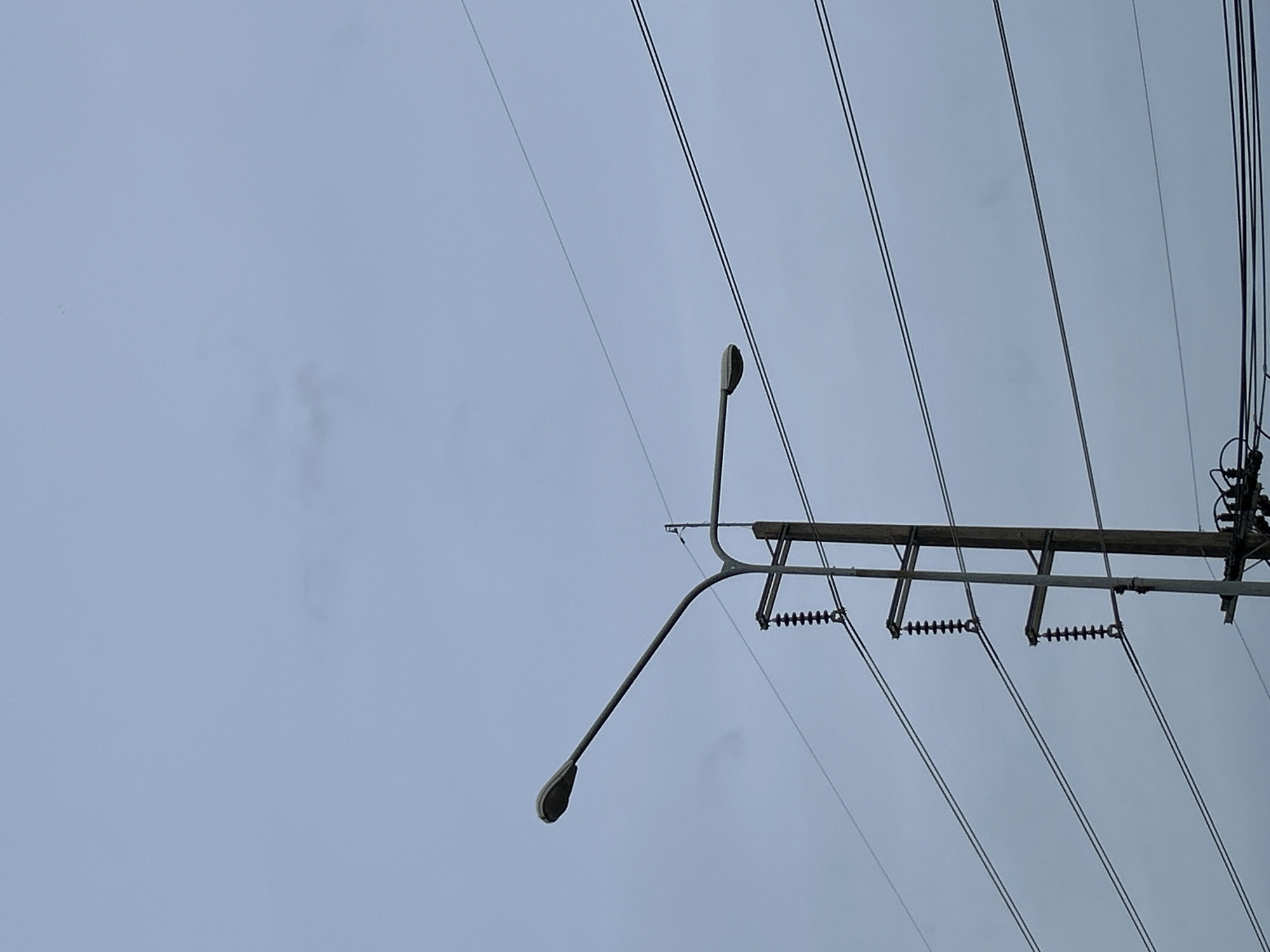 In Ratsada, Thailand, against a backdrop of a pale, overcast sky, a tall utility pole stands prominently. Extending from the pole is a light fixture with a curved, grey arm and a grey, oval-shaped light. Several dark electrical wires crisscross the frame horizontally. The utility pole itself is grey and appears to be made of metal. It has black insulators with multiple ribbed sections mounted on its side. The overall scene is stark and industrial, with the muted colors of the sky and the utilitarian nature of the pole and wires dominating the composition.