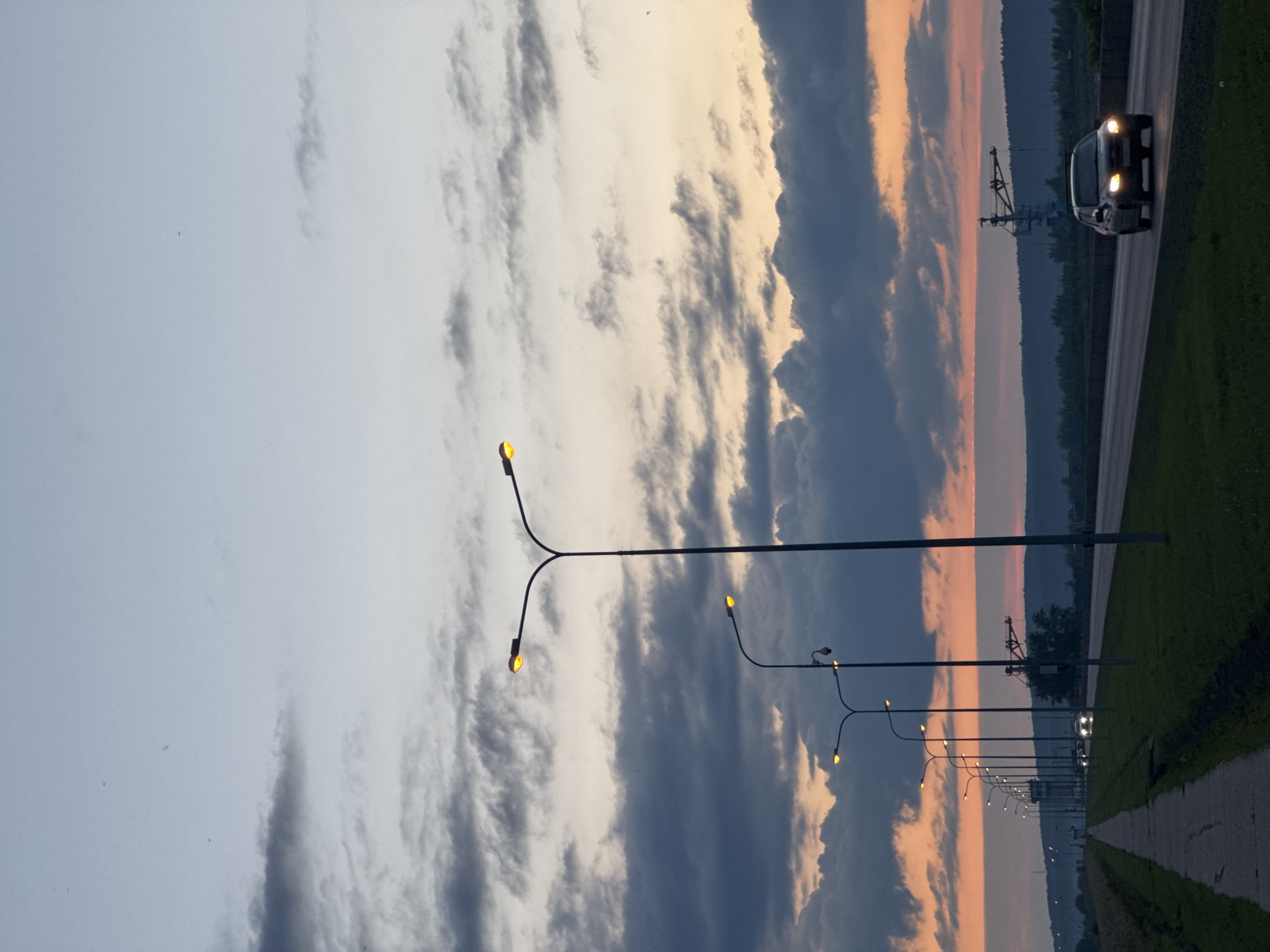 The image depicts a street scene at dusk in Chaikovsky, Russia. The sky is filled with dramatic clouds, painted with the warm hues of sunset in shades of orange and pink, contrasting with darker, grey clouds. A line of streetlights, illuminated with warm yellow light, stretches along the side of a road.
In the distance, a car with its headlights on is driving on the road, which is bordered by a grassy area and a paved walkway. The silhouette of hills and trees can be seen against the colorful sky. A notable object in the background appears to be some sort of industrial crane or structure. The overall mood is tranquil and serene, characteristic of the end of the day. The lighting suggests it is twilight. There is no visible text in the image.