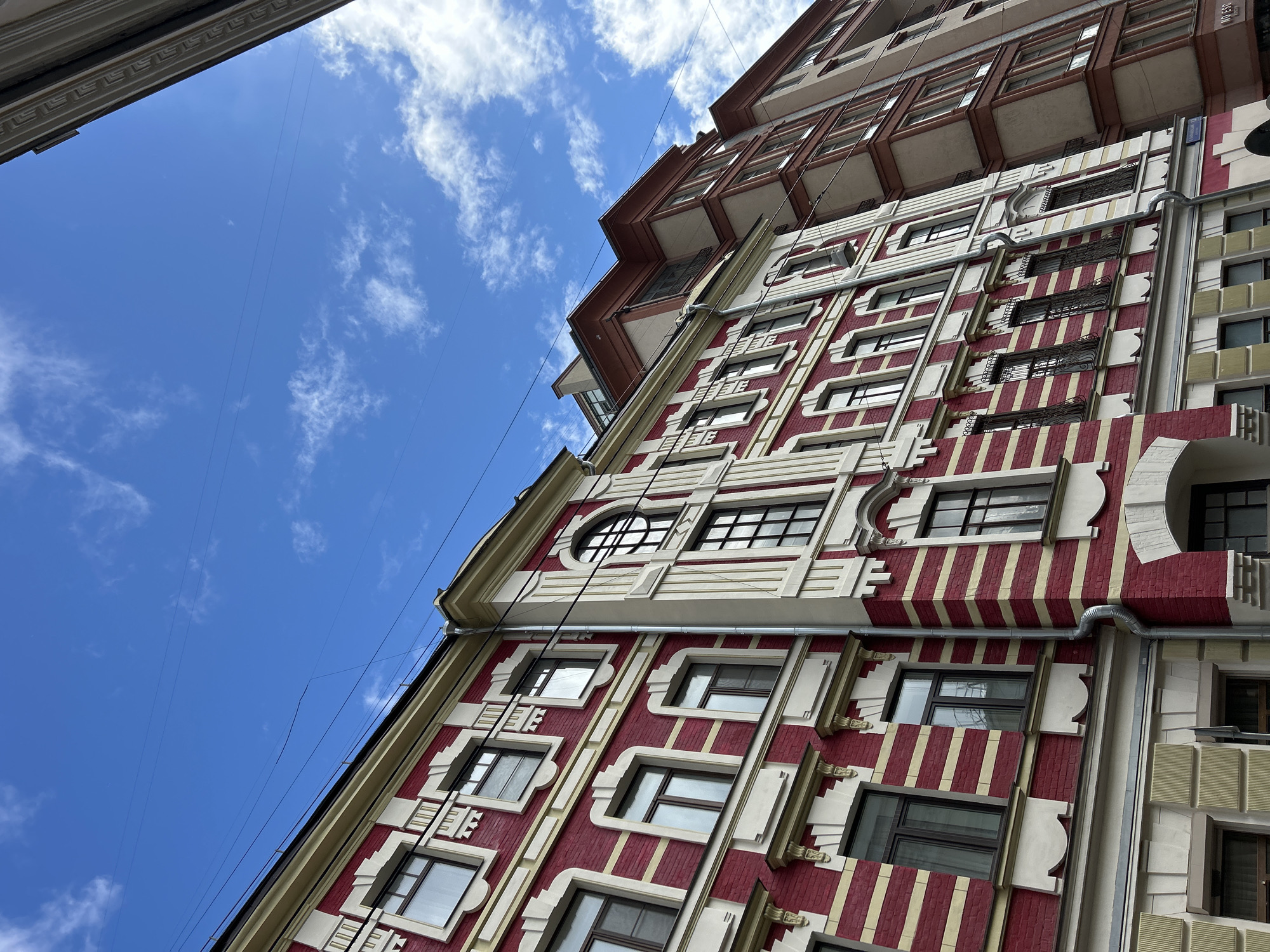 From a low angle, a multi-story building with red and white vertical stripes is visible against a blue sky with scattered clouds. The building has numerous windows with dark frames, surrounded by white geometric patterns. A large, round window is positioned near the top corner of the building. Gray drainpipes run vertically along the building's facade. Another building with brown and beige architecture is visible above and slightly behind the striped building. Power lines are strung across the sky in the upper-left corner of the frame. The location is Unknown, Unknown.