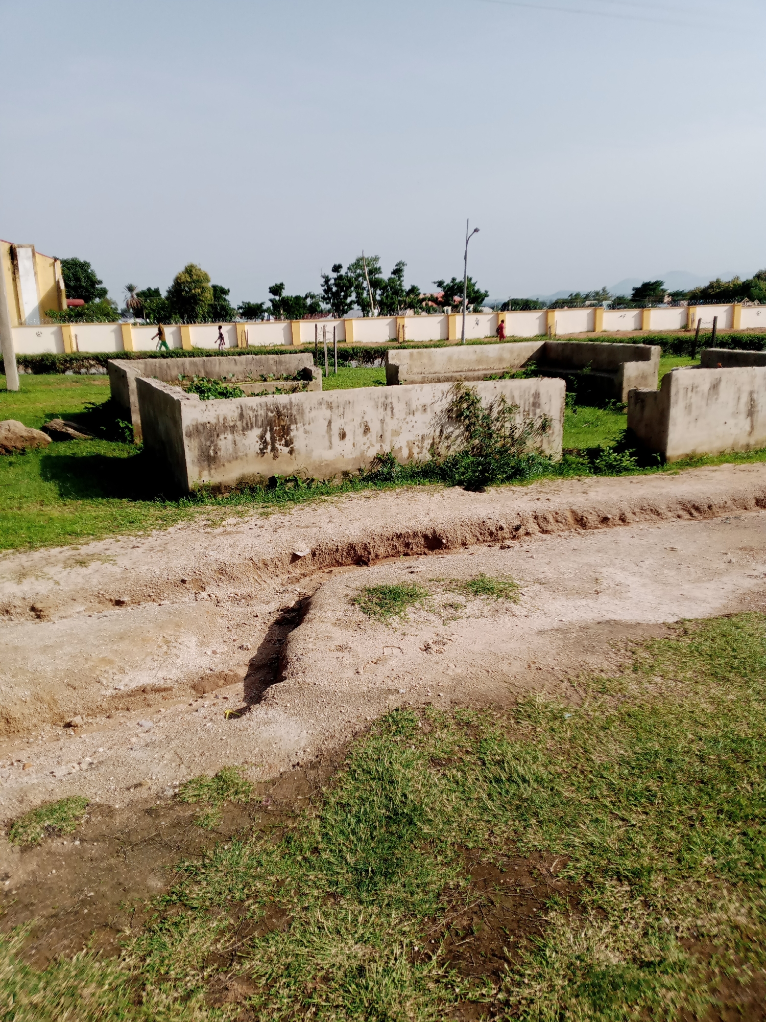 The image shows an outdoor scene in Jalingo, Nigeria. In the foreground, there is a dirt path with a trench dug into it. To the left of the path is green grass. In the middle ground, there are several concrete structures, possibly foundations or raised garden beds. Some of these structures have vegetation growing in or around them. In the background, there is a long, light-colored wall topped with barbed wire. Beyond the wall, there are trees and a clear sky. Two small figures, appearing to be children, are visible walking along the top of the wall in the distance. The overall impression is of an undeveloped or construction area.