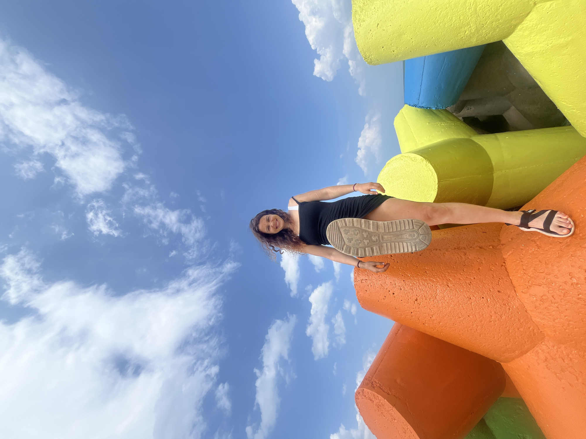 A young woman with curly brown hair is standing on brightly colored, concrete tetrapods against a blue sky with white clouds. She is wearing a black slip dress and dark sandals, and her right foot is raised, showing the sole of her shoe. The tetrapods are predominantly orange, yellow, and blue, arranged in a cluster that appears to form a breakwater or barrier. The perspective is from a low angle, looking up at the woman and the sky. The scene suggests an outdoor environment, likely a coastline or harbor area. It is not possible to determine the specific location within Phú Thủy, Vietnam based solely on this image.
