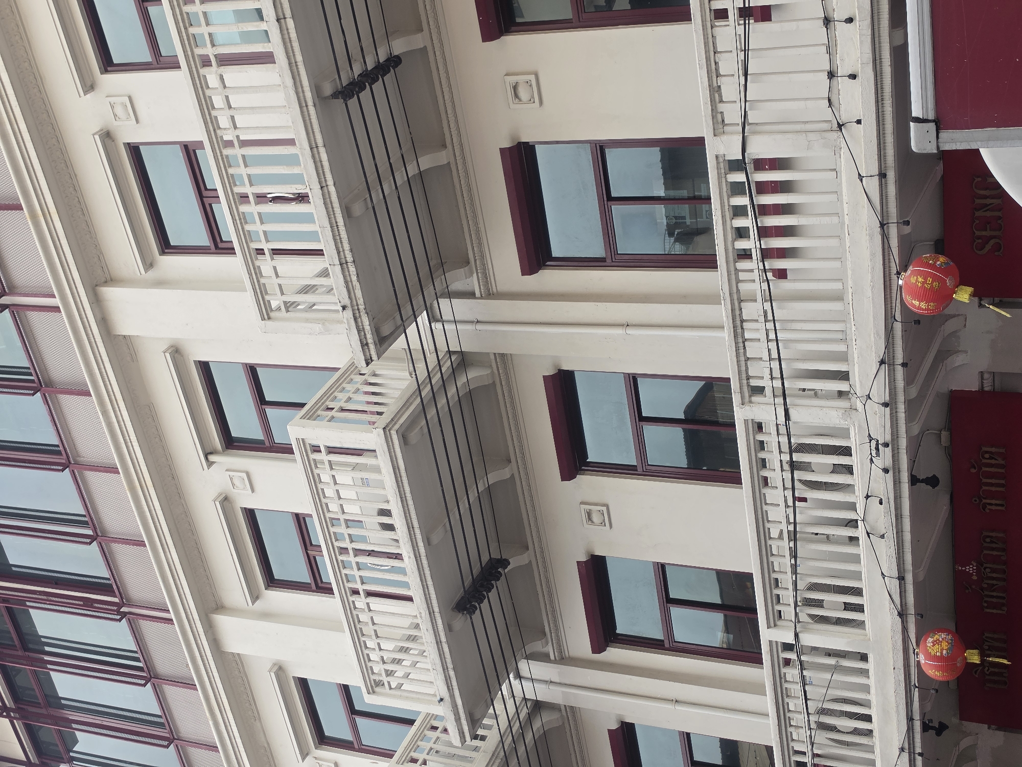 The image captures a section of a building in Bangkok, Thailand, displaying a blend of architectural styles and cultural elements. The building features multiple stories, distinguished by rows of windows with maroon frames and balconies with white balustrades. Some of the balconies have air conditioning units visible through the railings. Decorative elements such as ornate molding and small square plaques embellish the building's facade. Black electrical cables run vertically across the building. A red sign with Thai script and red Chinese lanterns are also visible.