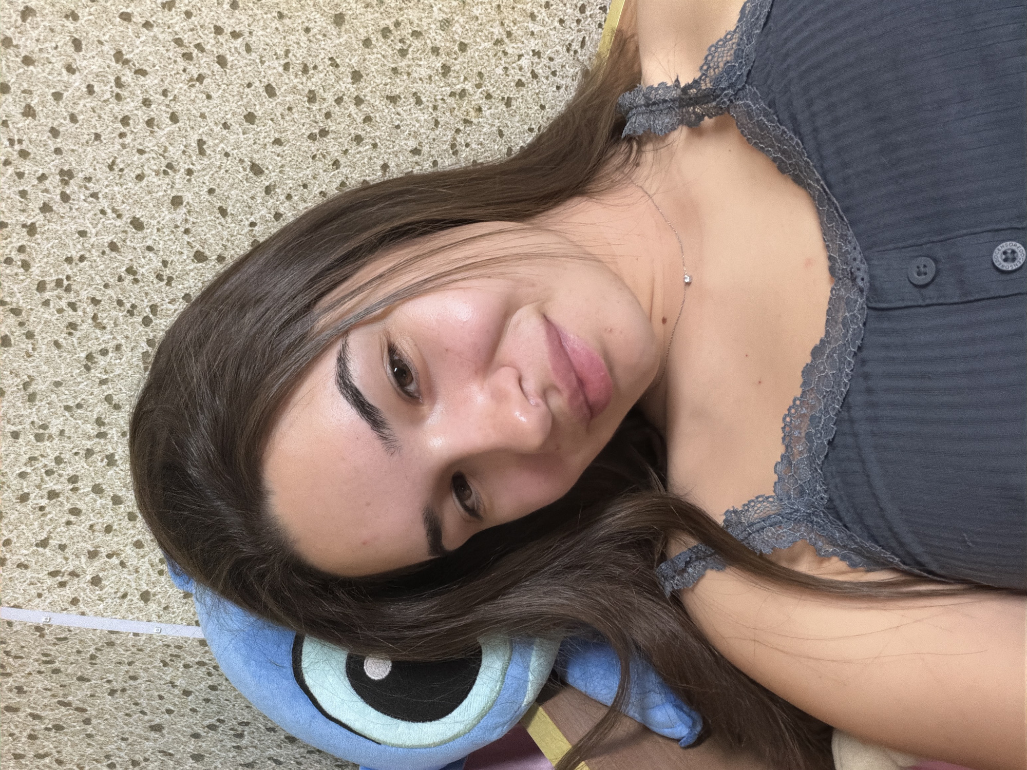 A woman lies on a patterned surface, her head resting on a blue stuffed toy with a large eye. She has long, dark hair and is wearing a gray tank top with lace trim. A delicate necklace adorns her neck. The background is a textured surface. The location is not evident in the image, and there is no indication that the photo was taken in Sofia, Bulgaria.