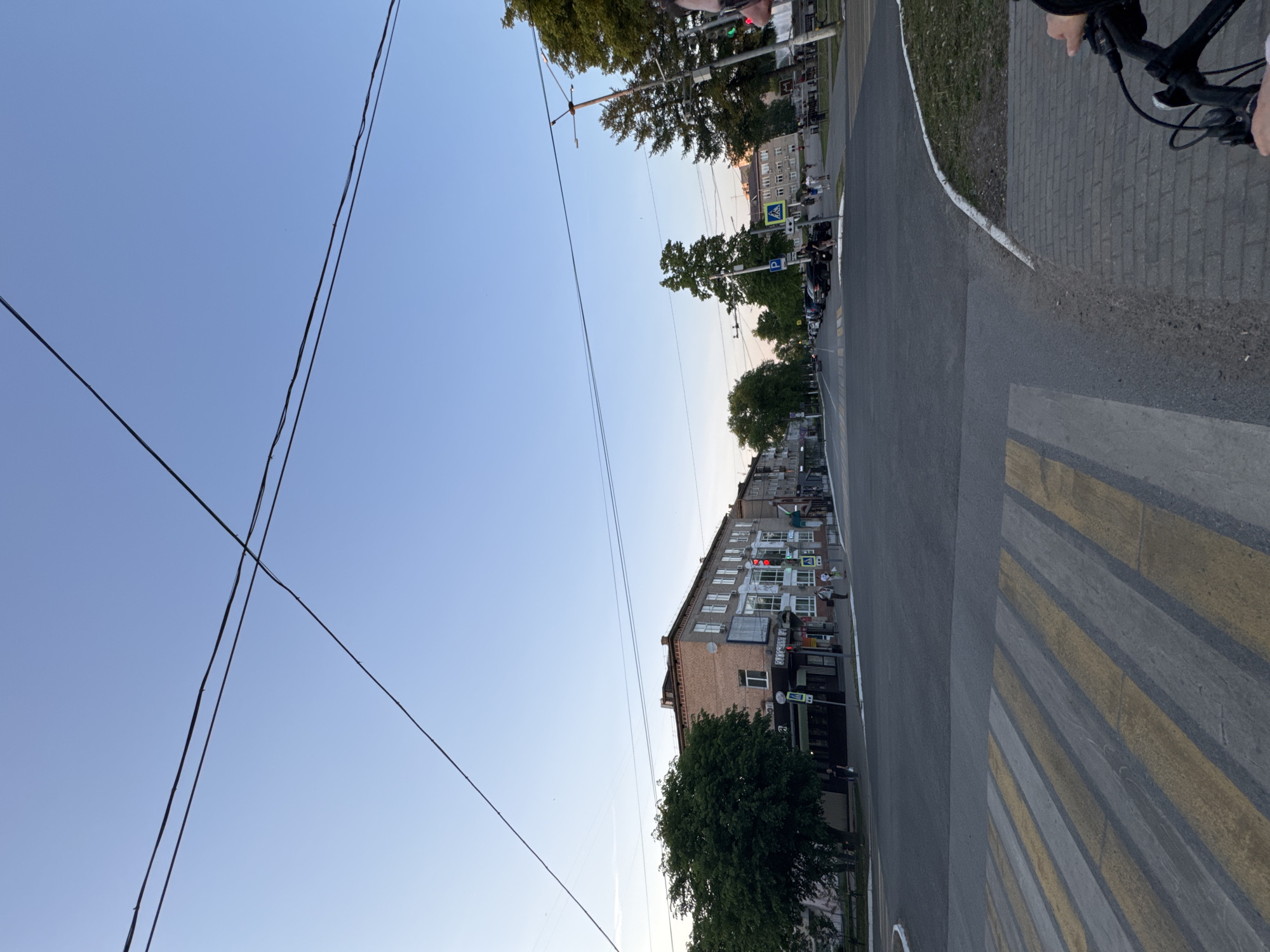 The image depicts a street scene in what appears to be a residential area. The camera is positioned on the edge of a street, possibly from a cyclist's perspective as a bicycle handle is visible in the top right corner. The street extends into the distance, flanked by buildings. On the right side, a sidewalk borders the street. The street is marked by a pedestrian crosswalk with yellow and gray stripes. Power lines intersect the sky. In the distance, there are pedestrians walking on the sidewalk. Buildings line the street, including a multi-story brick building with balconies. Trees are scattered along the street, partly obscuring some of the buildings. The sky is a clear gradient of blue, suggesting either dawn or dusk.