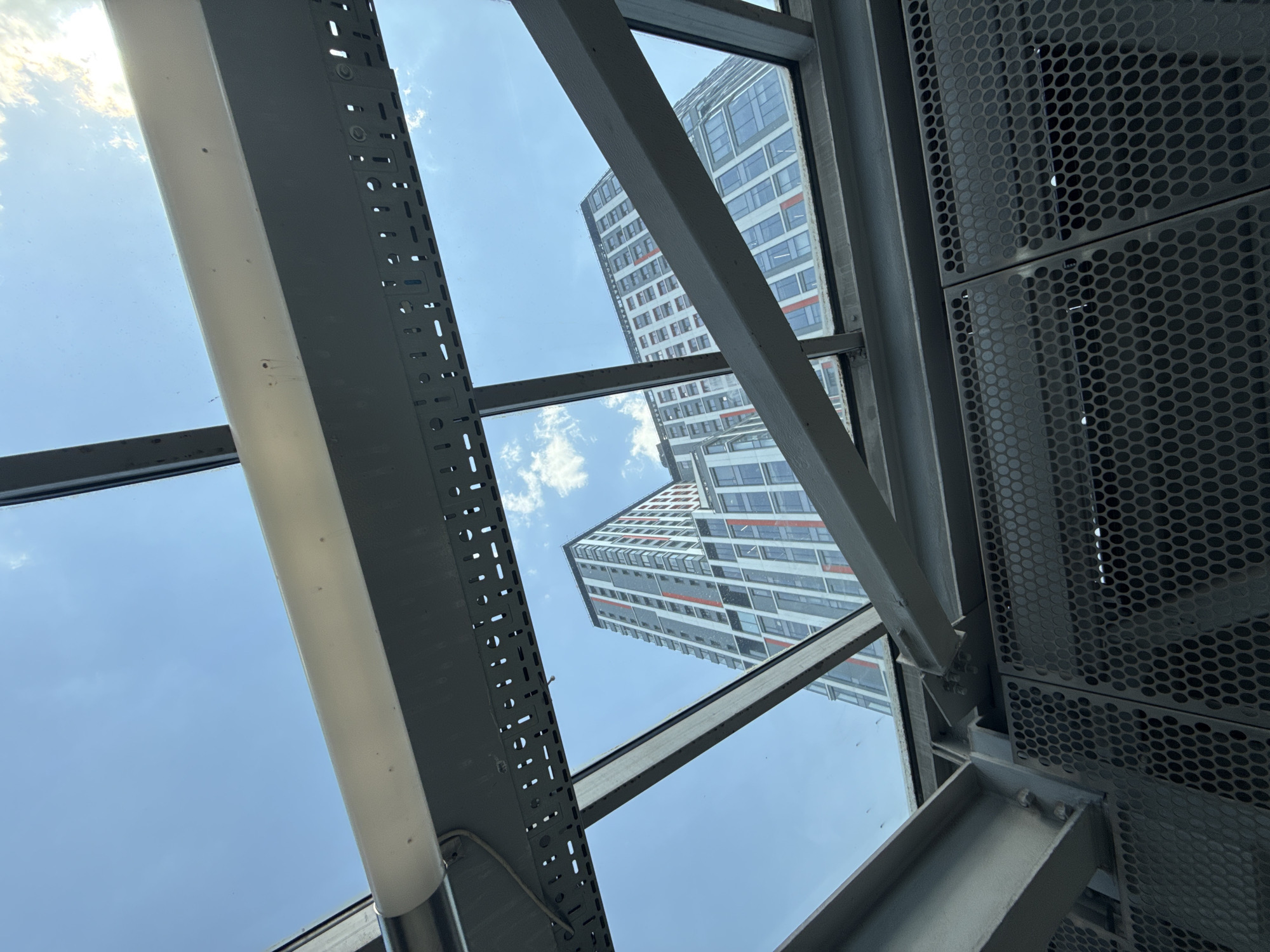 The image shows a view upwards through a structural framework, possibly a glass ceiling or canopy, in what is likely a public space. Two tall buildings are visible through the glass panels, partially obscured by the framework. The framework consists of metal beams and perforated metal panels. The sky is blue with a few scattered clouds. The buildings appear to be residential or office towers with repetitive window patterns and some orange accent stripes. Given the urban architectural style, the image is likely taken in a large city, and the location in the city of Moscow, Russia is based on the design and building styles observed in the image. There are no people visible in the image.