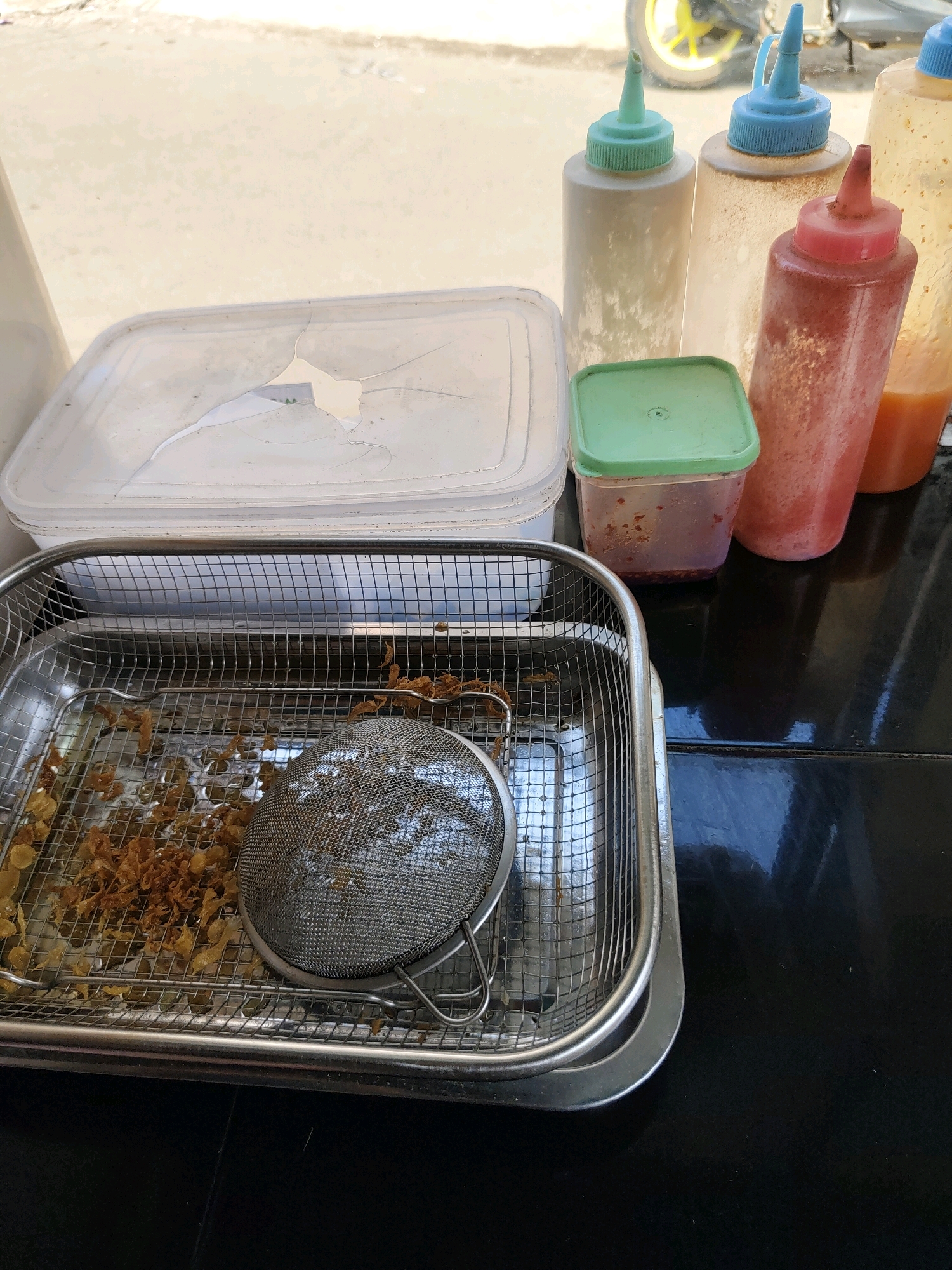 The image depicts a close-up, eye-level view of a food preparation area, likely outdoors in a city like Sukanagara, Indonesia. The foreground is dominated by a stainless steel rectangular basket filled with fried shallots and a small metal strainer. The basket sits on a black, reflective surface, possibly a table. Behind the basket, a white plastic container with a cracked lid is visible. Further back, several squeeze bottles are arranged, containing various sauces and condiments, their caps in different colors including green, blue, red, and a light orange. A small square plastic container with a green lid is also present between the white container and the squeeze bottles. In the background, out of focus, a motorcycle wheel with a yellow rim is visible, suggesting a street-side or open-air setting. The overall lighting is bright and natural, indicative of daytime.