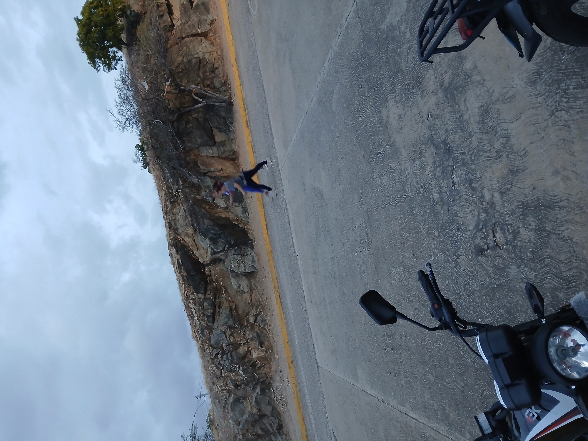 The image shows a scene on a road next to a rocky hill, likely in or near Juan Griego, Venezuela. A person is lying on the road near the edge, possibly performing a yoga pose or stretching. Parts of a motorcycle are visible in the foreground, including the handlebars, headlight, and front wheel. The road surface is paved, with a yellow line marking the edge. The sky is overcast. The environment consists of a rocky hillside with sparse vegetation.