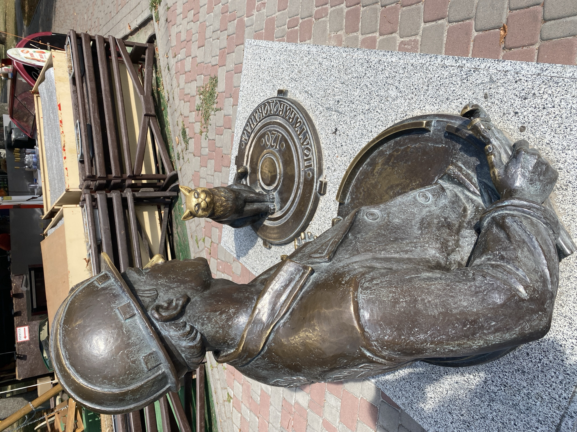 This is a medium shot, eye-level, outdoor photograph of a bronze sculpture in Poltava, Ukraine. The sculpture depicts a male figure wearing a hard hat and a jacket, seated on a granite base. Next to him, also on the granite base, is a bronze sculpture of a cat sitting on a manhole cover. The manhole cover is inscribed with Cyrillic text and the number "120". The background shows a paved walkway made of red and grey bricks, and behind that, wooden structures and a food stand. The lighting suggests it is daytime with the sun casting shadows. The sculpture appears to be a public monument.