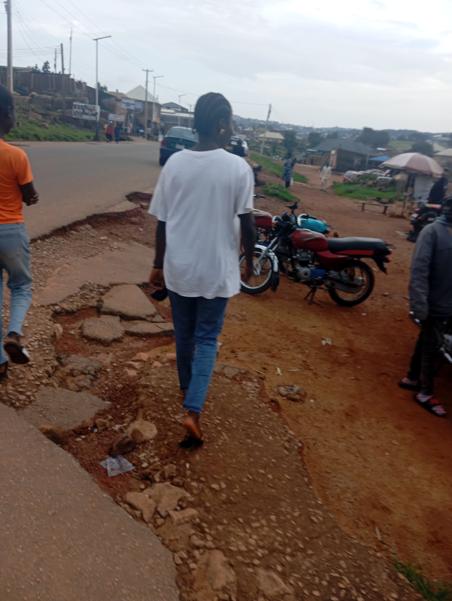 The image depicts a street scene in Jos, Nigeria, characterized by a rough, uneven asphalt road with significant erosion and exposed dirt. Two individuals are walking away from the camera on the left side of the road. The person in the foreground is wearing a white t-shirt and blue jeans, while the person further back wears an orange t-shirt and blue jeans. A red and silver motorcycle is parked on the right side of the road, with another motorcycle visible behind it. In the background, buildings and a few scattered trees are visible under an overcast sky. The environment appears to be a local, somewhat undeveloped area.