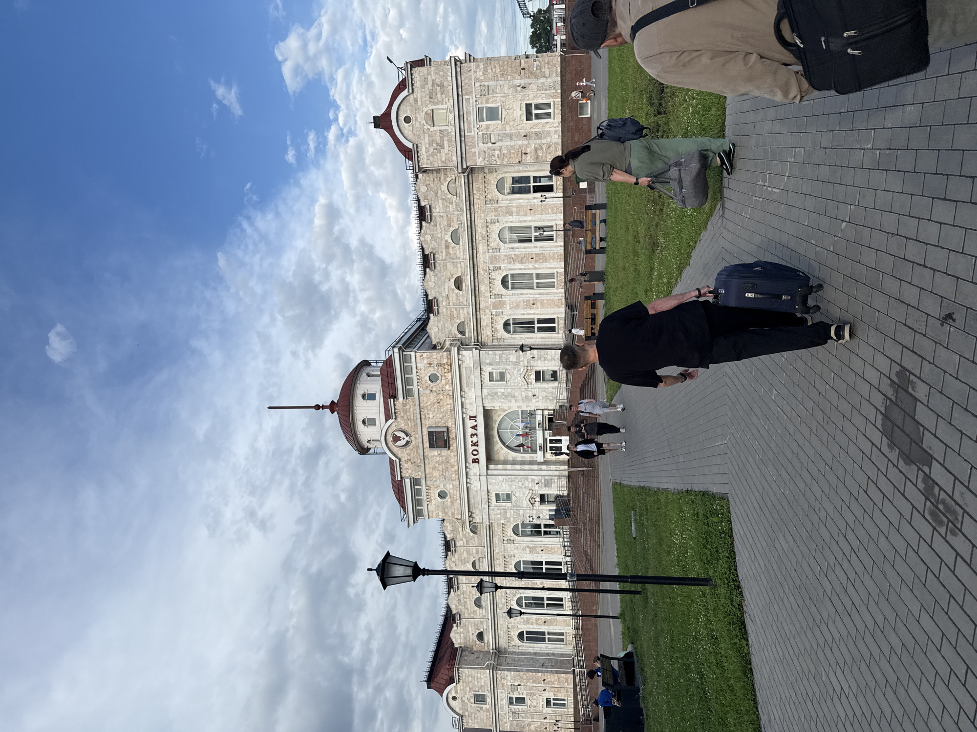 The image shows the Izhevsk railway station in Izhevsk, Russia, on a cloudy day. A man in a black t-shirt and black pants is walking on a paved pathway, pulling a blue suitcase. To his right, a woman in a green t-shirt and green pants is walking with a grey duffel bag. Behind her, another man in a beige jacket and dark pants is walking away from the camera, carrying a black laptop bag. In the background, the Izhevsk railway station building is visible, with its distinctive architecture and the word "ВОКЗАЛ" (VOKZAL - Station) above the entrance. There are also lampposts, benches, and grass in the scene. Several other people are visible in the distance, including some sitting on benches and others walking. The sky is partly cloudy with patches of blue.