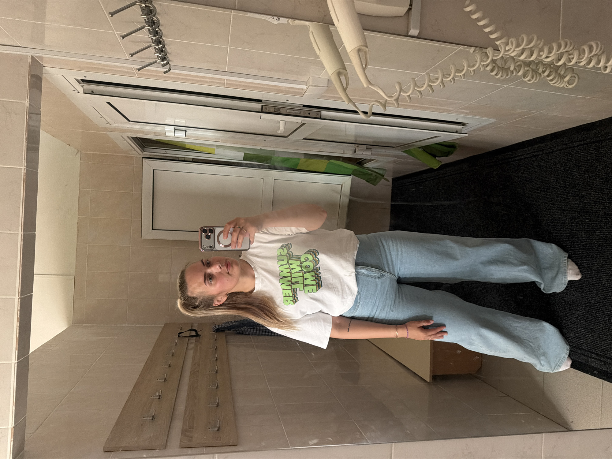 This is a vertical, mirrored selfie of a young woman in what appears to be a locker room or changing area. The woman has blonde hair pulled back in a ponytail and is wearing a white t-shirt with green text and light blue wide-leg jeans. She is holding a smartphone in her right hand, taking the photo. She is wearing rings on her fingers and bracelets on her left wrist.
The background shows tiled walls, a large mirror, and a white door with a metallic frame. Above the door, there are coat hooks. To the right of the door, there's a dark mat on the floor. In the reflection, we can see a white coiled cord, possibly from a hairdryer, hanging from the ceiling. The lighting suggests it is daytime indoors.
The text on her t-shirt is partially obscured and mirrored, but appears to say "COME WIT SUMMER" in a blocky, 3D font with a green gradient. The overall mood is casual and relaxed. Given the setting and attire, the woman is likely changing or has just finished changing. The location context of Chaikovsky, Russia is noted.