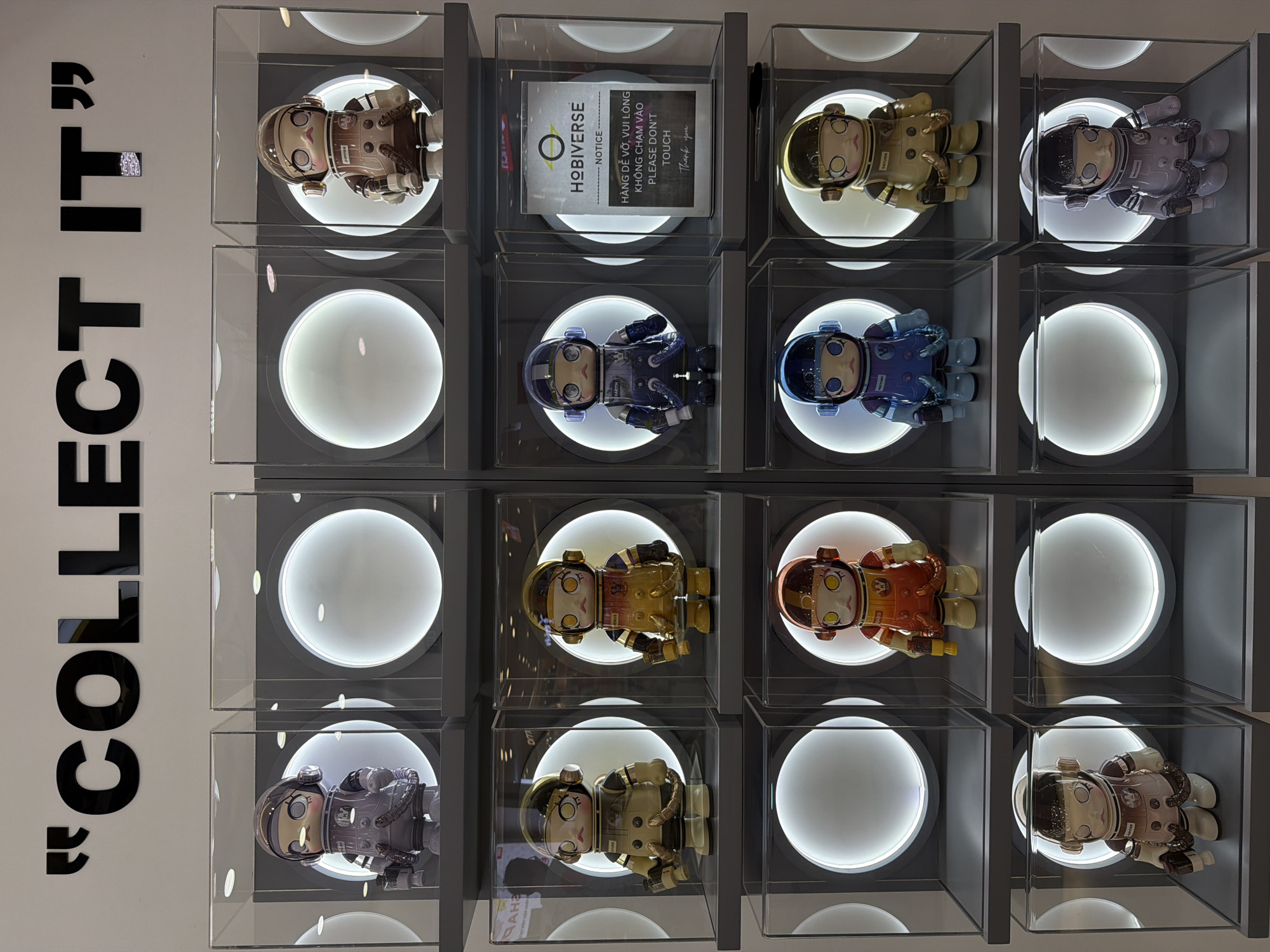 The image shows a display case with twelve compartments, each containing a figurine in an astronaut-like suit. The figurines vary in color and design. A sign in the center of the case reads "HOBIVERSE - NOTICE" and includes text in both English and Vietnamese, stating "PLEASE DON'T TOUCH" and "Thank you." To the left of the display, bold black lettering spells out "COLLECT IT" vertically. The overall setting suggests a retail or exhibition space in Đà Nẵng, Vietnam, as indicated by the Vietnamese text on the sign.
