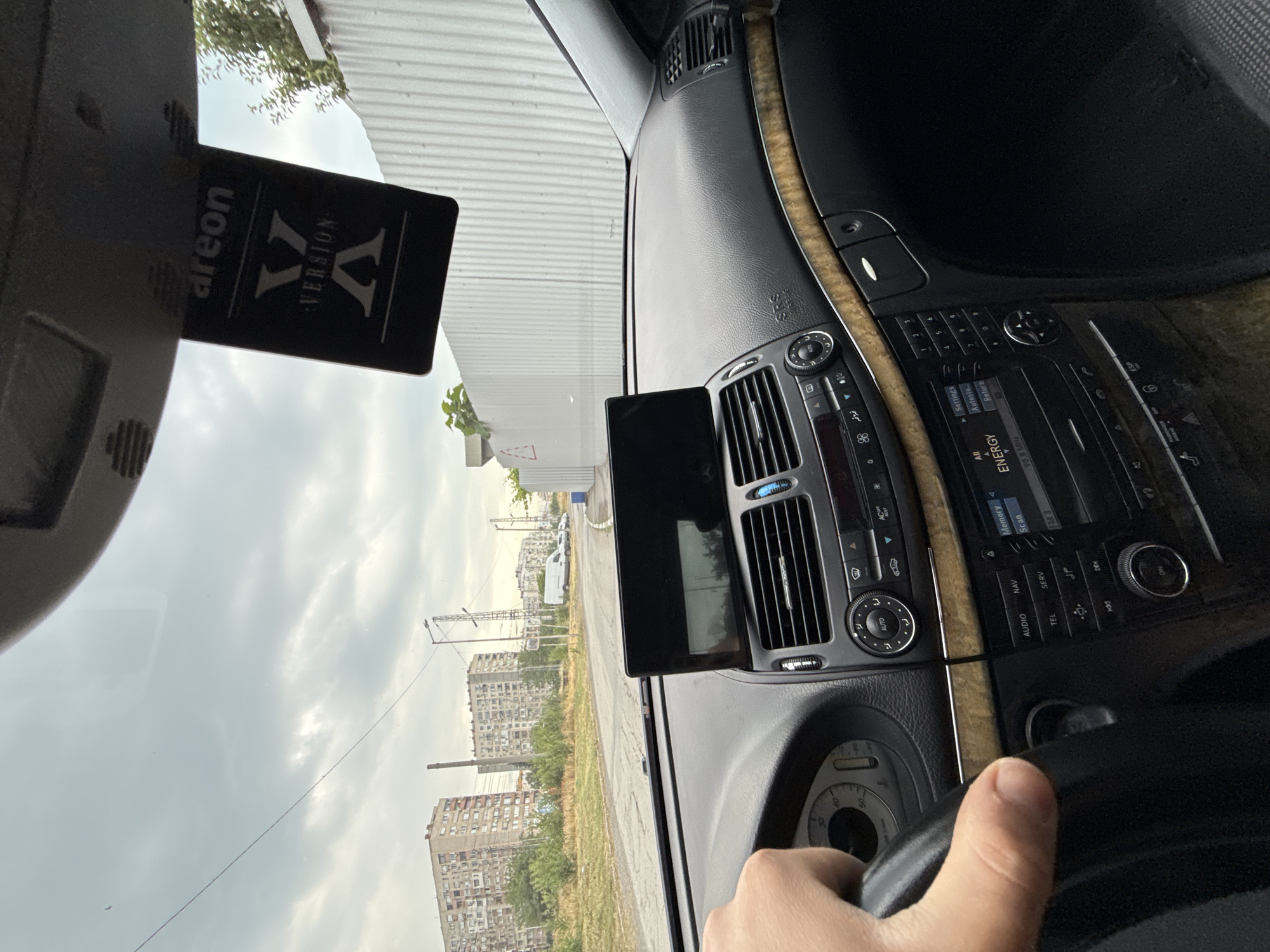 The image depicts the interior of a car, viewed from the driver's perspective through the windshield, looking out towards an urban landscape under an overcast sky. A human hand, likely the driver's, is visible in the foreground gripping the steering wheel on the right side of the frame.
Inside the car, a black dashboard with wood trim runs across the width of the vehicle. The central console features an infotainment system with various buttons and controls, including "AUTO", "ACOFF", "ESP", "NAV", "AUDIO", "TEL", and "SERV". Text on the display reads "ENERGY", "FM 3", "90.8 MHz", "Memory", "Scan", "Settings", "Autosto", and "Return". An airbag indicator displaying "SRS" is also visible. A black rectangular electronic device, possibly a phone or tablet, is mounted on the dashboard near the central air vents. From the rearview mirror hangs a black rectangular air freshener with the text "areon" and "VERSION: X". The speedometer is partially visible, showing numbers like "30", "40", and "50". The car appears to be a Mercedes-Benz, possibly an E-Class (W211 generation), judging by the interior design.
Outside the car, the scene reveals an urban environment consistent with Stara Zagora, Bulgaria. Several large, multi-story residential buildings, characteristic of post-socialist architecture, dominate the background. A factory chimney and utility poles are also visible. The ground appears dry with sparse vegetation, leading to a road. The sky is entirely covered with clouds, indicating an overcast daytime setting. The car seems to be parked or slowly moving through this urban area.