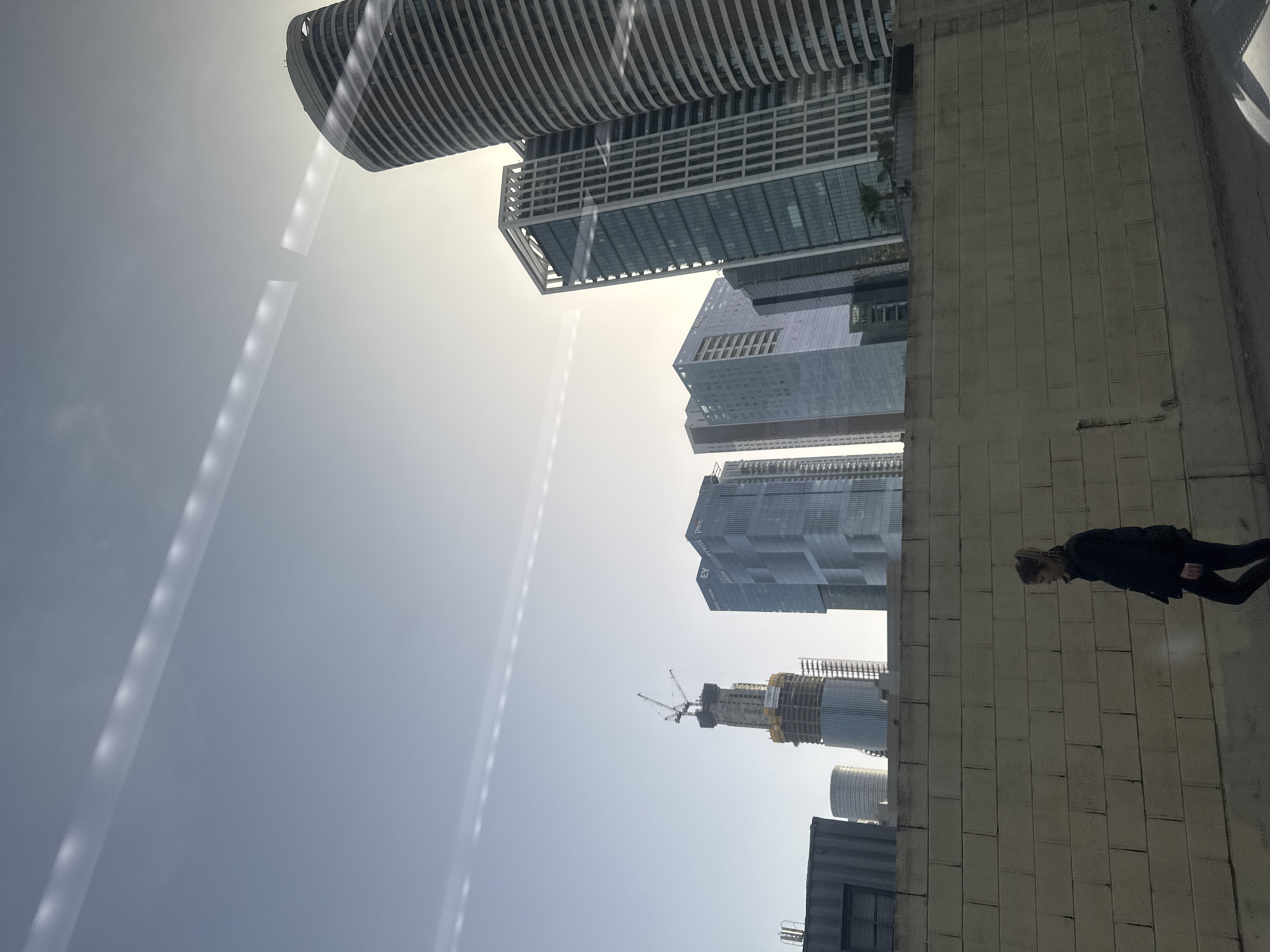 The image depicts a scene in Tel Aviv, Israel. It shows a person in a dark coat, walking along a concrete structure that appears to be a wall or building. In the background, there are several skyscrapers, including one under construction with a crane visible. The sky is overcast with streaks of light.