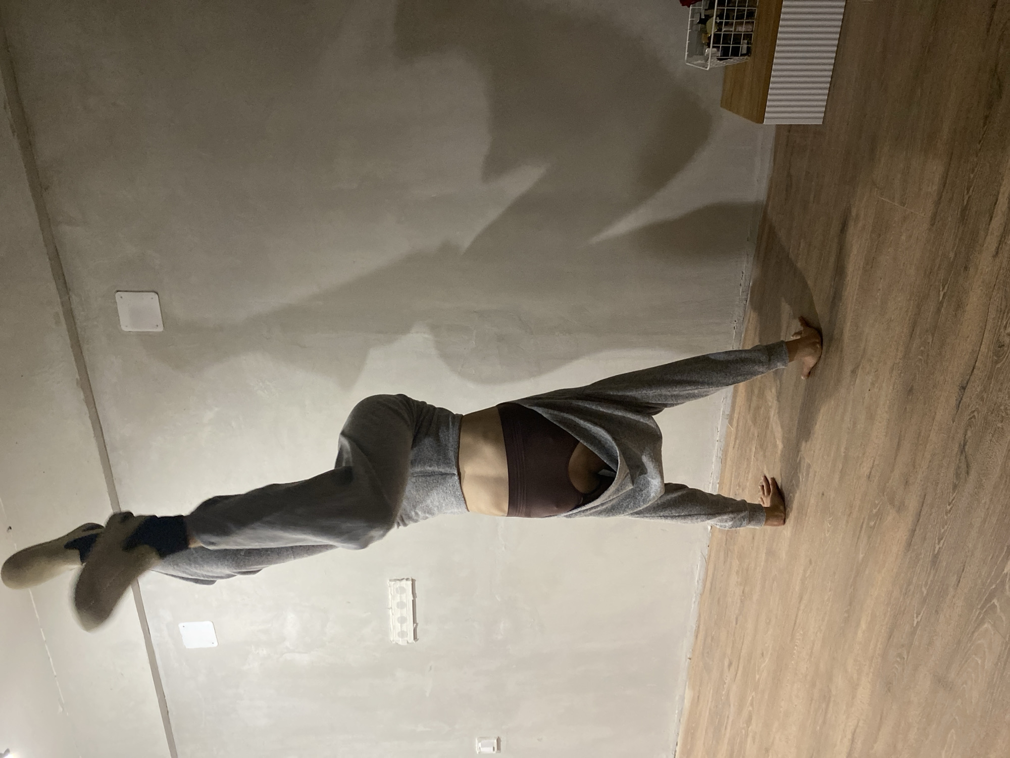 A person wearing gray sweatpants and a brown sports bra is performing a split between a gray wall and a wood-look floor. The wall has several small electrical outlets and a ceiling light. A piece of furniture and wire basket are against the wall on the right. The location is an interior space. The city of Votkinsk, Russia, is not evident in the image.
