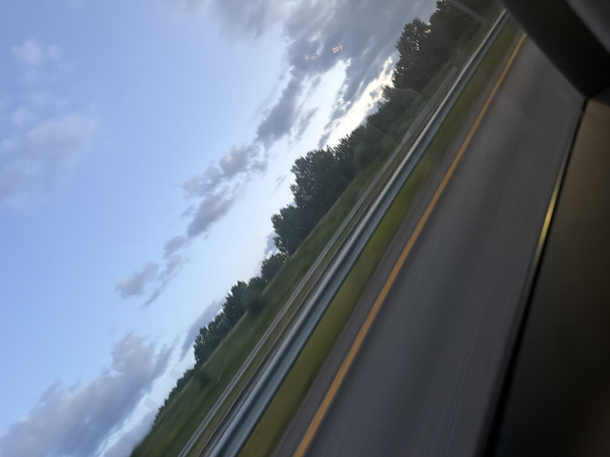 The image depicts a view from a moving vehicle, likely a car, looking out a window. The perspective is at an angle, suggesting the vehicle is traveling on a road, possibly a highway, as indicated by the presence of a yellow dividing line and a metal guardrail. The scene is characterized by motion blur, making sharp details indistinct.
The environment appears to be rural or semi-rural. On the left side of the road, a green, grassy embankment slopes upwards and is lined with a dense row of dark green trees. Beyond the trees, the sky is visible, a light blue with scattered, wispy clouds, some of which are tinged with gray, hinting at dusk or early morning. The overall lighting suggests it is not midday.
There are no people or animals visible in the image. The primary subjects are the road, the guardrail, the vegetation, and the sky. The motion blur is a significant characteristic, indicating the photograph was taken while in transit.
Regarding the location in the city of Sarpsborg, Norway: without specific landmarks or identifying features, it is impossible to pinpoint the exact location within Sarpsborg. However, the scene is consistent with a road in a more developed area that transitions into or borders natural landscapes, which is common in and around many Norwegian cities. The presence of a highway with a guardrail and tree-lined embankment is a general characteristic that could be found in various parts of Sarpsborg.