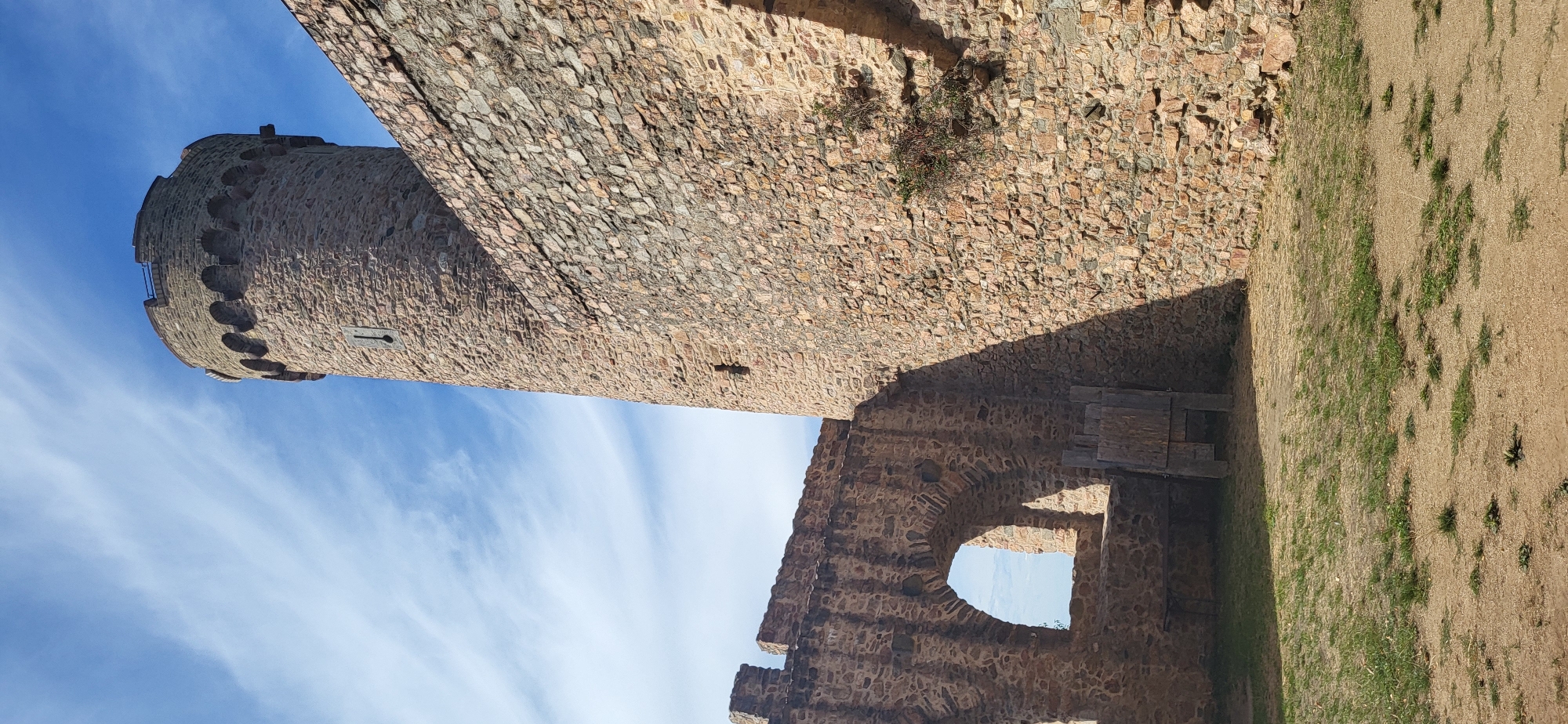 The image depicts a view of a stone tower and a section of a stone wall, with an arched window opening in the foreground. The tower, which is round and tall, is constructed of rough-hewn stones and topped with battlements. A narrow slit window is visible on its side. Adjacent to the tower is a segment of a thick stone wall, also built with irregular stones, showing signs of age and some vegetation growing on its surface. In front of the wall is a large, arched window opening that looks out onto a blue sky with wispy white clouds. The ground in the foreground is a mix of sandy soil and patchy grass. To the left of the arched window, there are some wooden structures that appear to be lean-to or support beams. The scene appears to be part of a historical ruin or a preserved castle. This is a view of the Auerbacher Schloss, a castle located on the Schlossberg above the town of Auerbach, a district of Bensheim, Germany.
