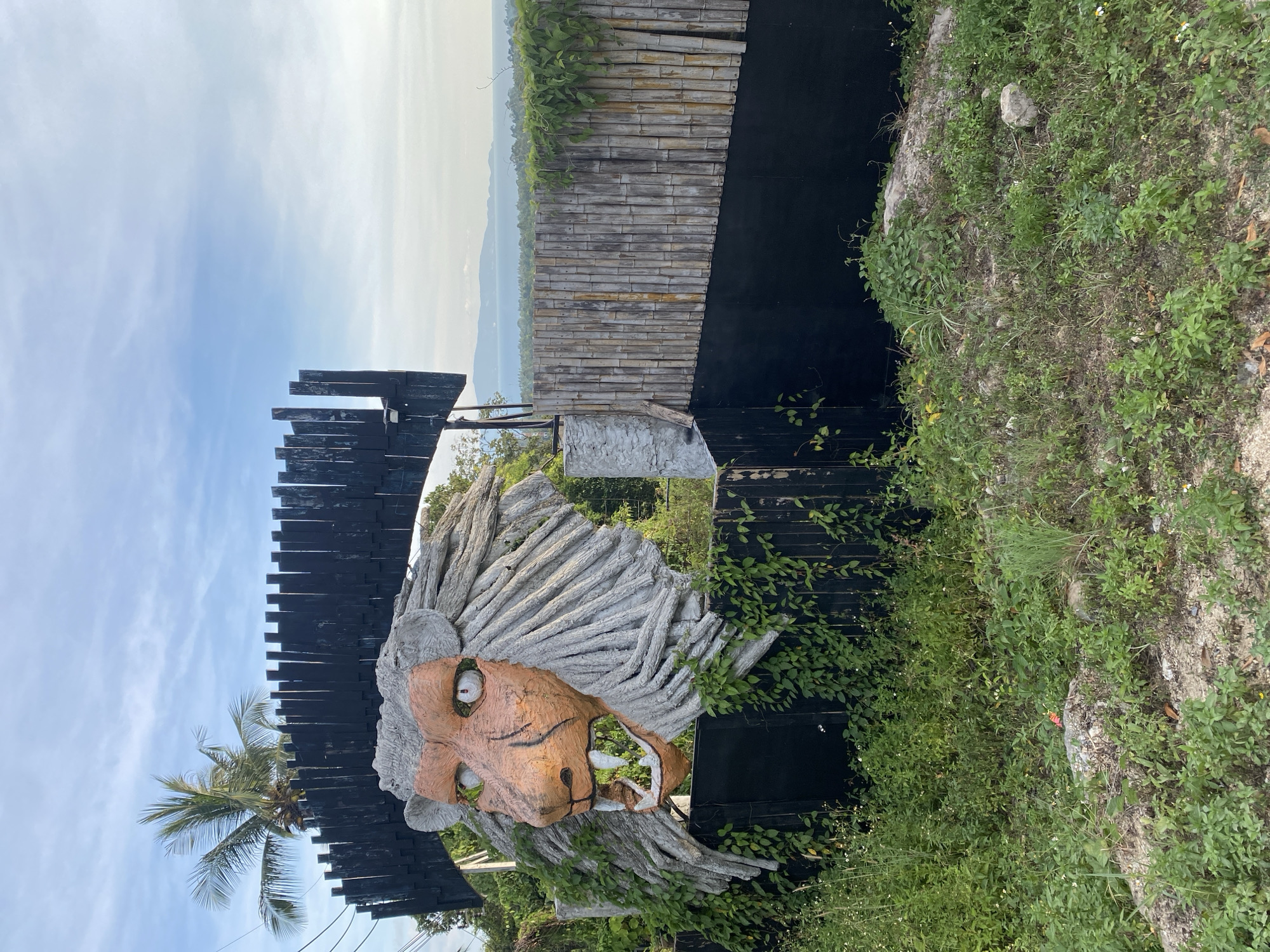 A large lion head sculpture sits at the entrance to a building in Moo 2, Thailand. The sculpture is constructed from gray concrete-like material, with a tan-colored face, green eyes, and prominent white teeth. Its mane is made up of numerous concrete strands. The sculpture is mounted on a black wooden structure. Behind the wooden structure, there's a wall made of bamboo. The area surrounding the entrance is overgrown with green foliage and vegetation. The sky is partly cloudy.