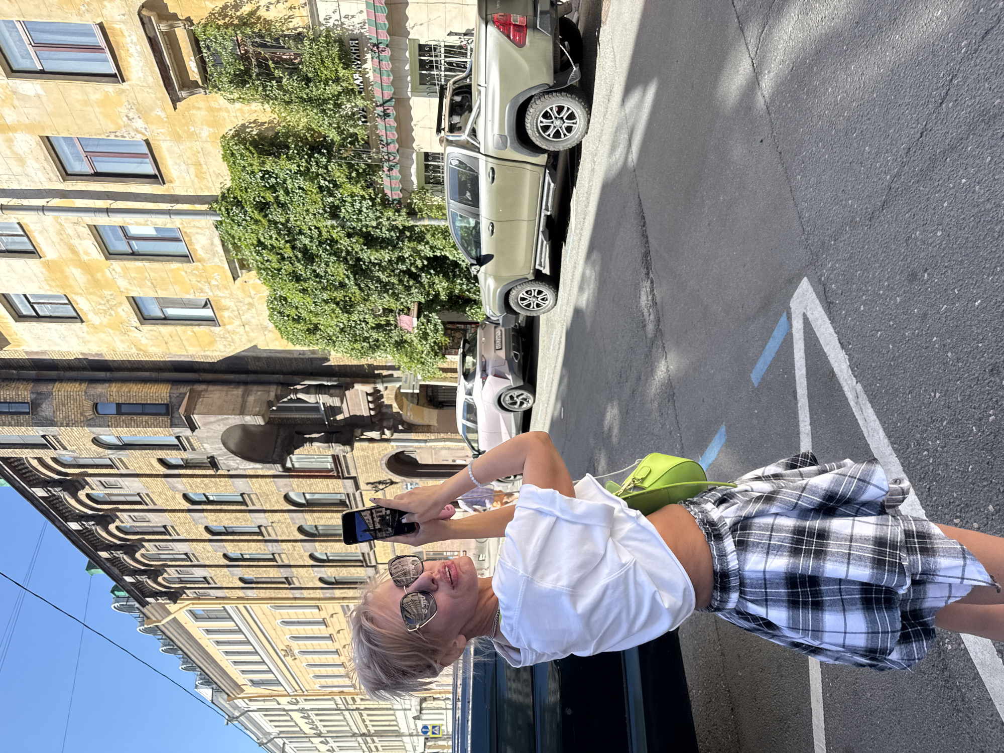 In a sun-drenched urban scene in Saint Petersburg, Russia, a young woman with blonde hair and sunglasses is captured from the waist up. She is holding a smartphone, taking a picture of herself or her surroundings. The woman is wearing a white, cropped t-shirt and a black and white plaid mini-skirt, accessorized with a vibrant lime green handbag slung over her shoulder. Her expression suggests a moment of joy or amusement.
Behind her, the scene unfolds with architectural details typical of Saint Petersburg. A pale yellow building with numerous windows of varying shapes and sizes forms the backdrop. Ivy crawls up the side of one of the buildings, adding a touch of nature to the urban landscape. In the middle ground, a light-colored pickup truck is parked on the street, its tires and details visible. Further back, another white car is partially visible. The street itself is paved asphalt, with white parking lines painted on it. The clear blue sky indicates a sunny day. The overall impression is of a warm, bright day in a historic European city, with a focus on a fashion-conscious individual enjoying a moment of leisure.