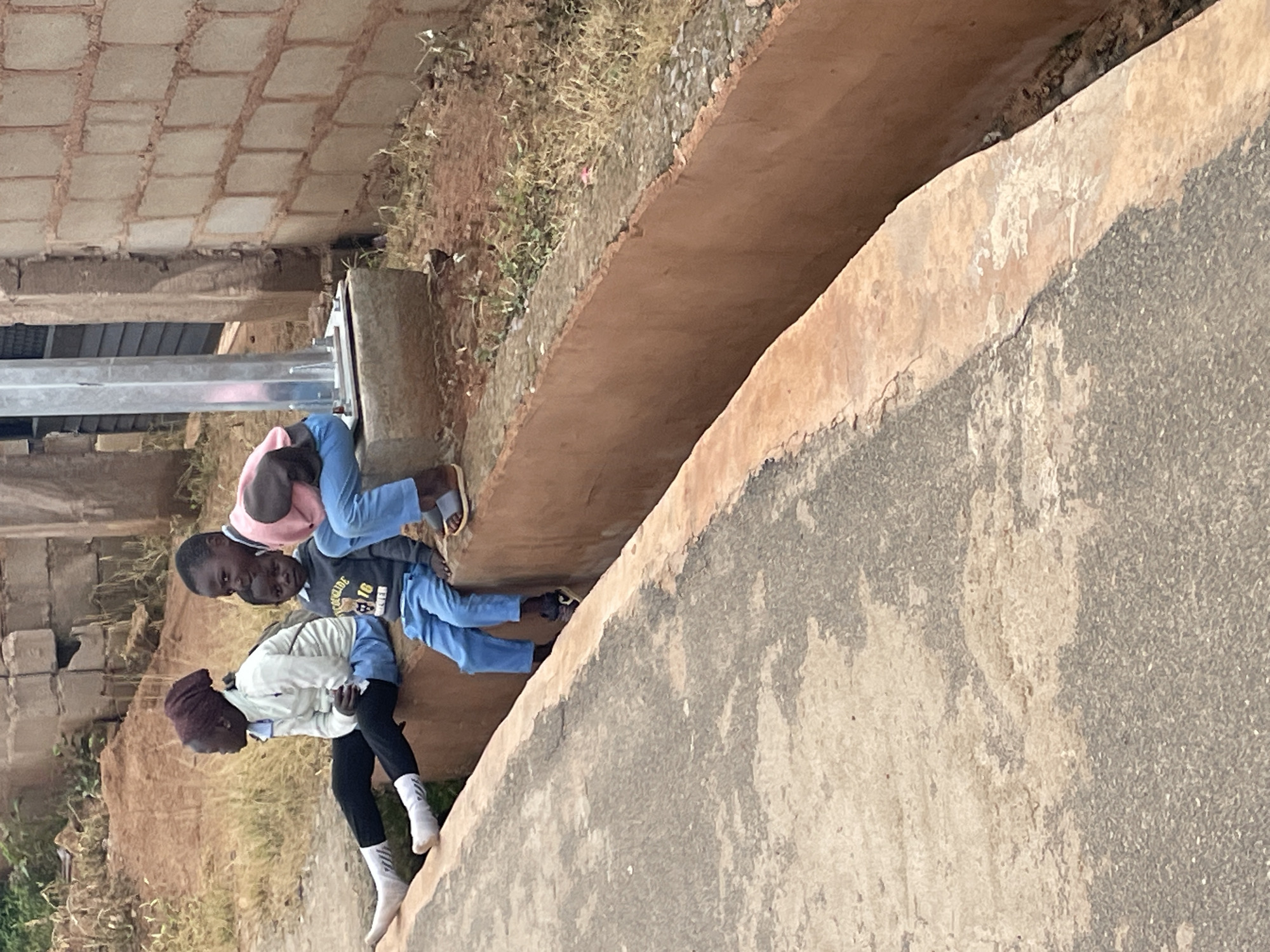Two young Black boys are sitting on a concrete structure in Gura Top, Nigeria. The boy on the left is wearing a white jacket, black pants, white socks, and a maroon head covering. He is leaning forward and looking down. The boy on the right is wearing a pink shirt and blue pants, and he is sitting upright. He is looking towards the camera. They appear to be in a rural or semi-rural setting, with dry grass and dirt visible around them. The concrete structure they are sitting on looks like a raised embankment or drainage channel. The image is taken outdoors, and the lighting suggests it is daytime, possibly late morning or early afternoon, with a slight overcast. There is no discernible text in the image.