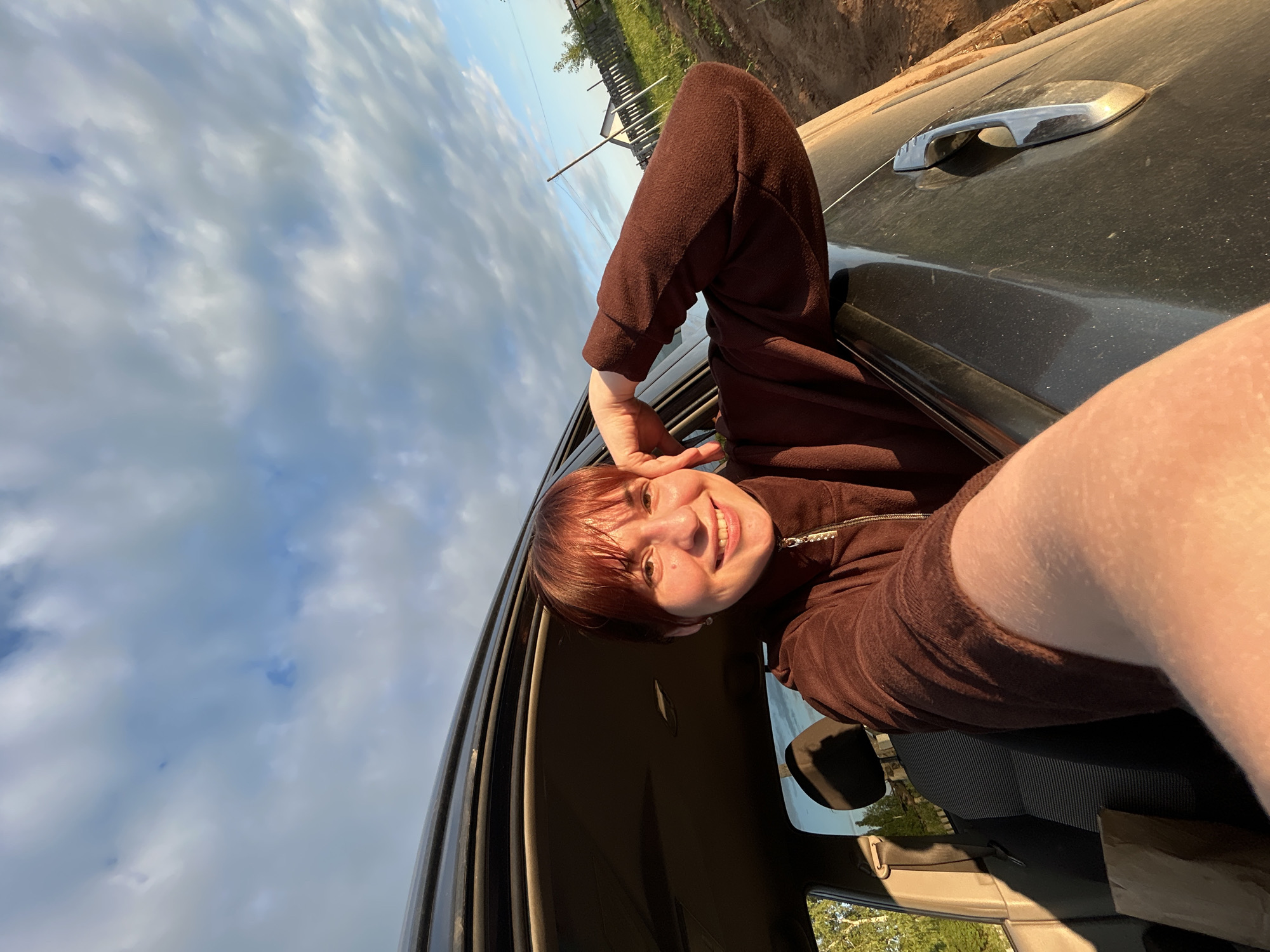 A joyful young person is captured leaning out of a dark-colored car's driver-side window. The image is taken from a low angle, looking up, and is rotated 90 degrees clockwise. The person has short, reddish-brown hair with bangs and is wearing a brown, textured, long-sleeved top with a metallic zipper detail. They are smiling widely at the camera, showing teeth, and appear happy and relaxed.
The car's exterior, specifically the door and window frame, is visible, appearing dark and slightly dusty. A silver-colored car door handle is seen on the dusty door. Inside the car, a side mirror is partially visible.
The background features a bright, partly cloudy sky, with shades of light blue and white clouds dominating the upper portion of the frame. Towards the horizon, above the car, hints of a rural or semi-rural landscape are visible, including green vegetation, a wooden fence, and what appears to be a dirt road or embankment, characteristic of a setting like Фоки, Russia.
The lighting suggests late afternoon or early evening, with warm sunlight illuminating the person's face and the car's exterior, creating a pleasant glow. The overall impression is one of carefree enjoyment during a car journey. No visible text is present in the image.