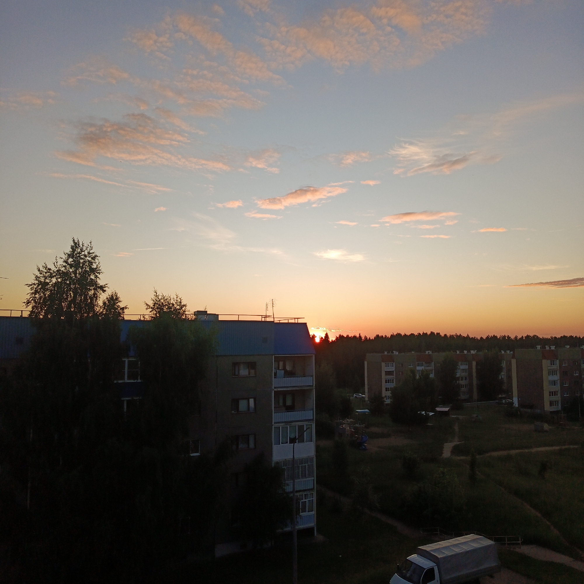 The image depicts a panoramic view of a residential area in Markovskiy, Russia, during either sunrise or sunset. The sky transitions from a pale blue at the zenith to a vibrant orange and yellow hue near the horizon, with scattered, elongated cirrus and altocumulus clouds displaying pink and light orange coloration. The sun, a bright orange disc, is partially obscured by the dark silhouette of a dense coniferous forest that forms a distinct line across the midground horizon.
In the immediate left foreground, a multi-story apartment building with a corrugated blue and grey roof and multiple balconies is partially visible, largely obscured by several dark, deciduous trees. Antennas are mounted on its roof. To the right of this building, a smaller, darker structure is visible behind more trees.
Extending across the midground, behind the forest line, are several additional multi-story apartment buildings characterized by light-colored facades (likely beige or light brown) and darker, possibly red-tiled roofs. Faint outlines of vehicles are visible parked near these buildings.
The lower right section of the image features a grassy, unkempt area traversed by winding, unpaved dirt paths. A children's playground structure with a blue slide is partially discernible in the midground between buildings. In the bottom right corner, a white light truck with a grey canvas bed cover is parked, facing towards the left. No individuals are visibly present in the scene.