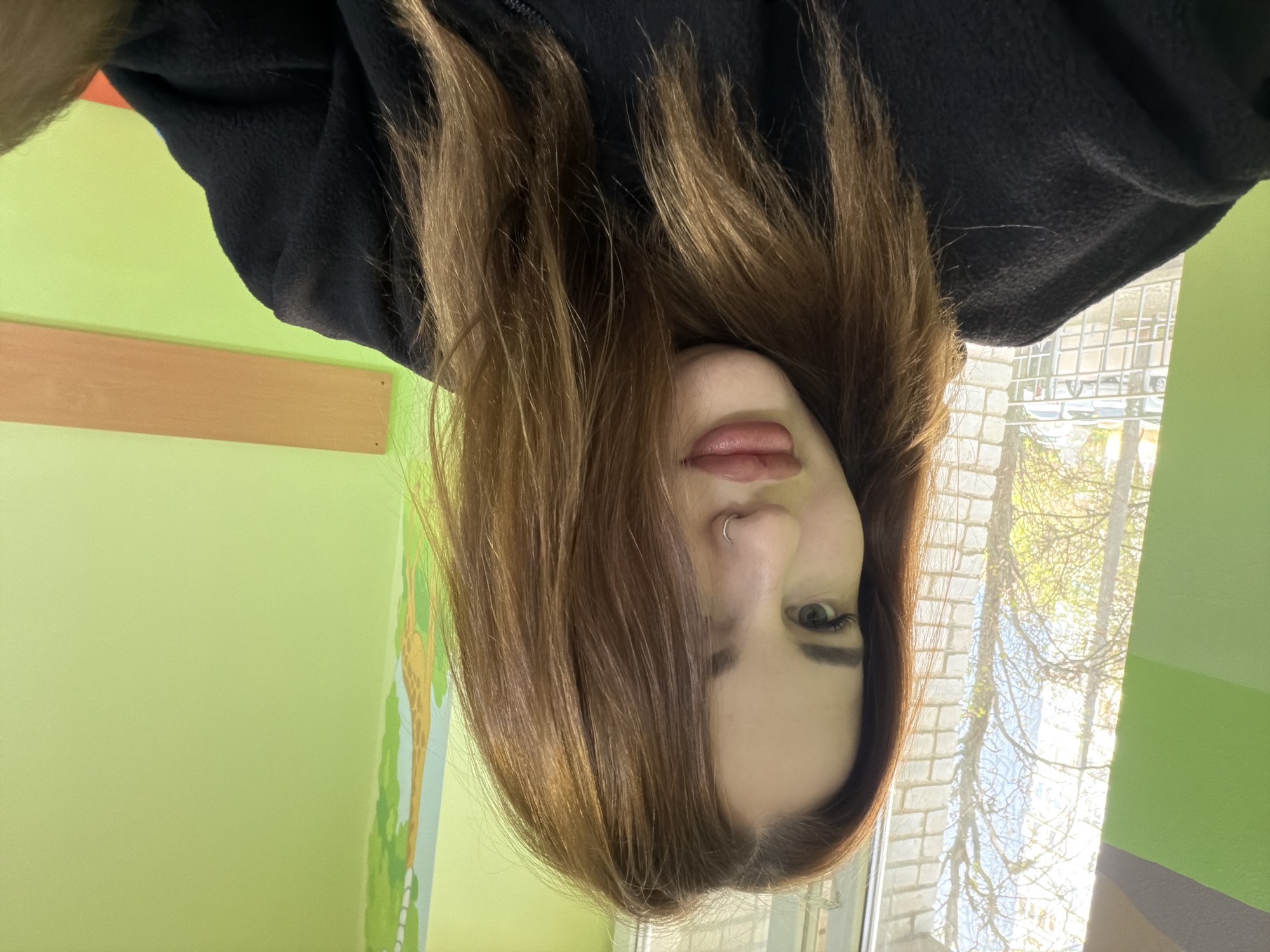 A person is shown in a selfie-style image, which has been rotated 180 degrees. The person has brown hair, a nose ring, and is wearing a black top. The background includes a green wall with what appears to be a mural or design on it, and a window showing a brick building and trees outside. The location is Unknown, Unknown.