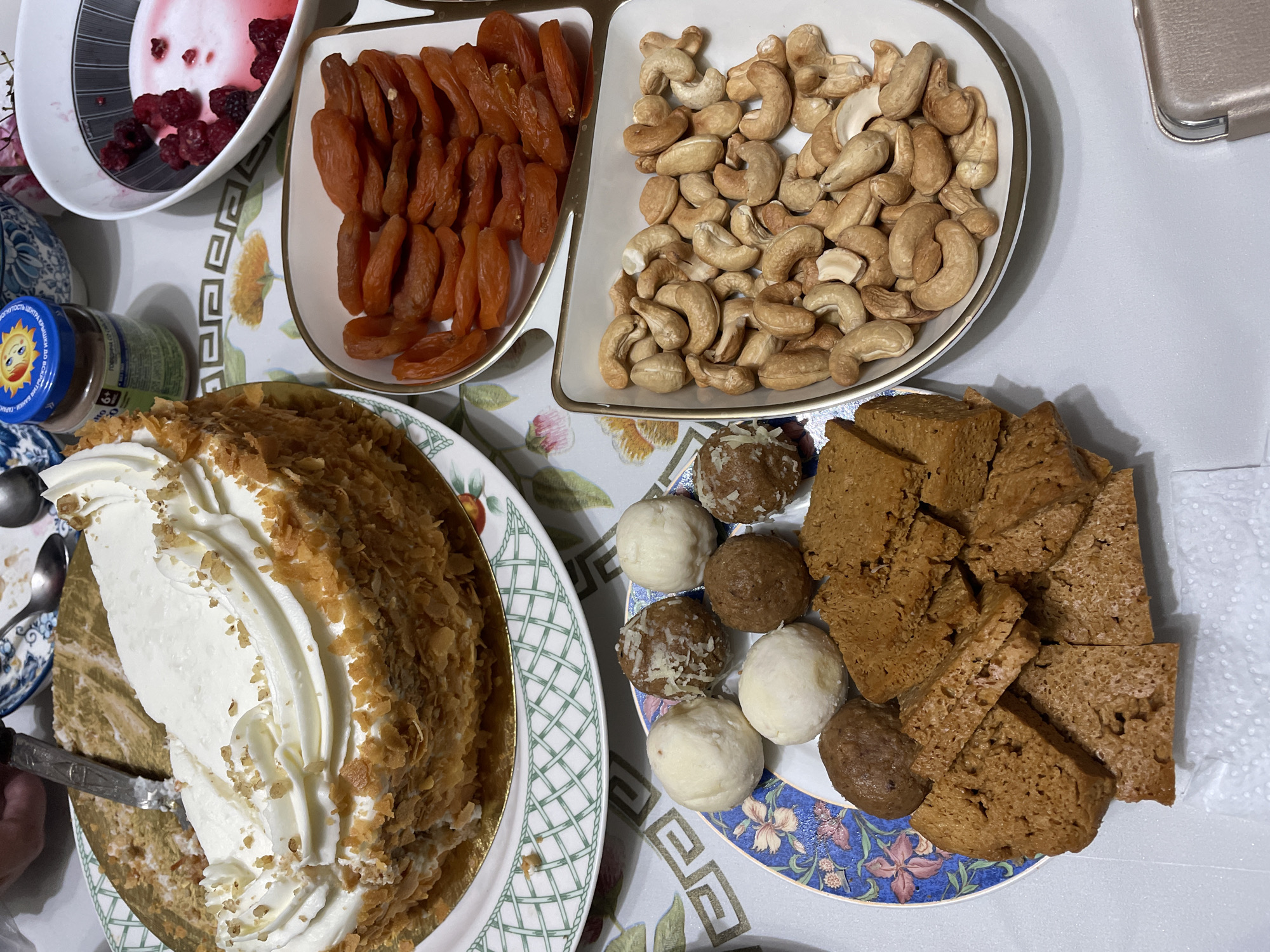 A collection of sweets and dried fruits is laid out on a table, suggesting a gathering or celebration. A large, layered cake, generously topped with whipped cream and crushed nuts, takes center stage. Beside it, a plate holds a variety of small, round candies, some appearing to be chocolate-based and others white. Another plate displays an assortment of dried apricots, neatly arranged. Adjacent to the apricots, a bowl is filled with a generous portion of cashew nuts. In the upper left corner, a small bowl contains a pinkish dessert with visible berries, possibly raspberries. A jar with a blue lid and a smiling sun graphic is also visible. The setting appears to be indoors, with a patterned tablecloth underneath the food. The overall impression is one of a dessert spread, likely for a special occasion.