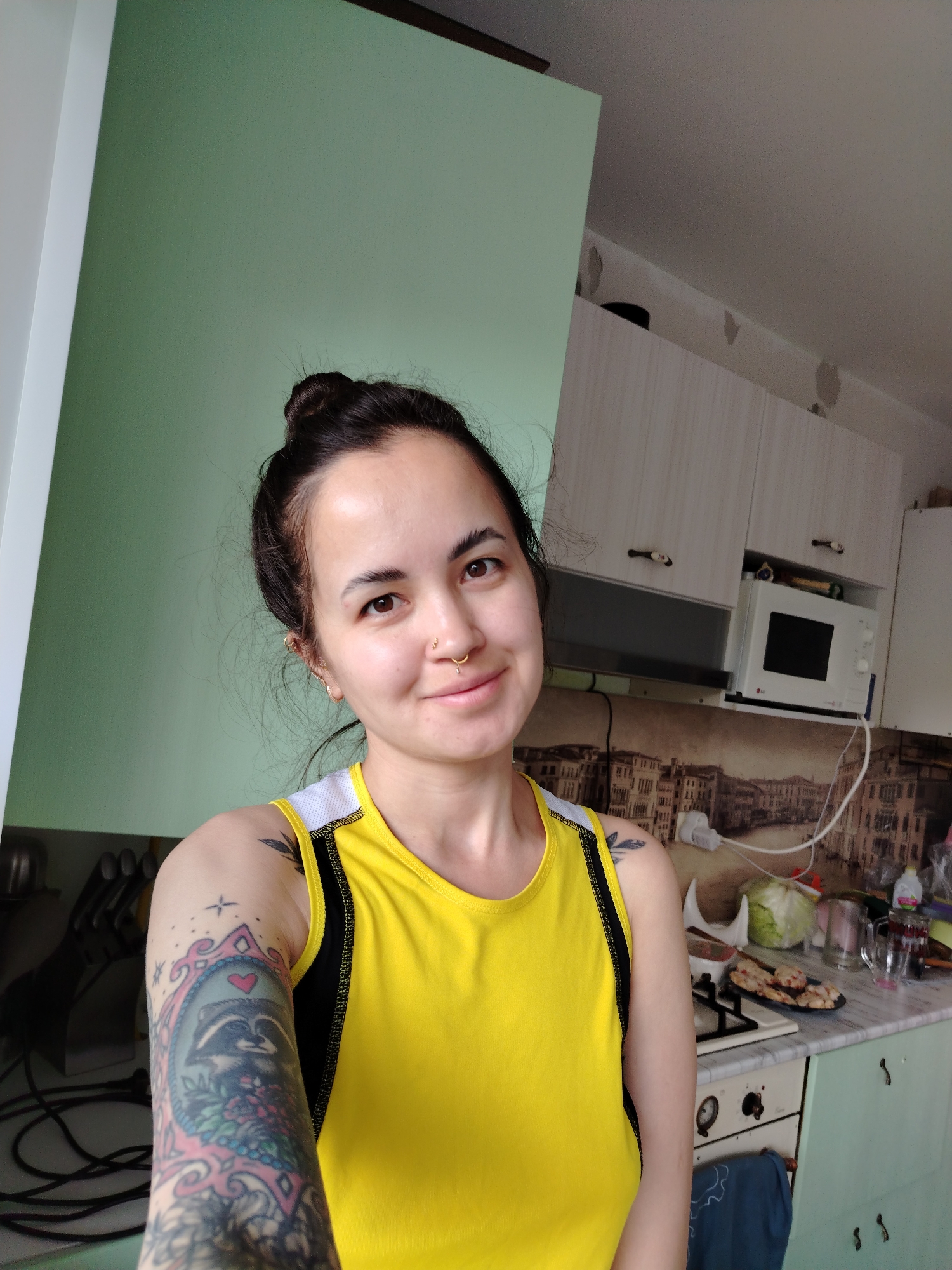 A woman is taking a selfie in a kitchen. She has dark hair pulled back in a bun, a nose ring, and tattoos on her left arm. She is wearing a yellow tank top with black and white trim. The kitchen has light green walls and white cabinets. There is a microwave on the counter and a cityscape backsplash behind the stove. There appears to be food on the stove. The woman's ethnicity is unclear. Based on the location, it may be Noviy, Russia, or another place with a similar name.