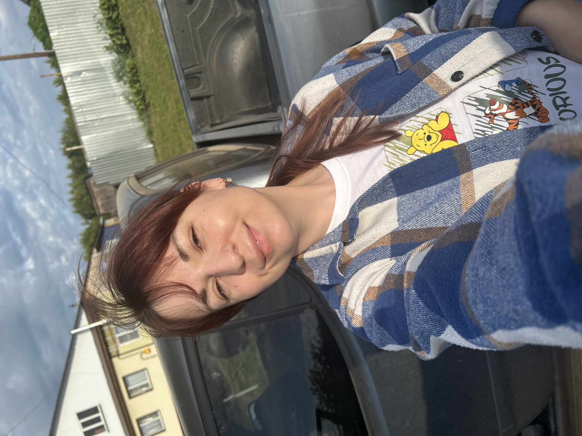 A woman with red hair and a white shirt featuring Winnie the Pooh and Tigger is wearing a blue and brown plaid flannel shirt and leaning on a dark-colored vehicle. The background shows buildings and trees under a partly cloudy sky. The image was taken in Fokino, Russia.