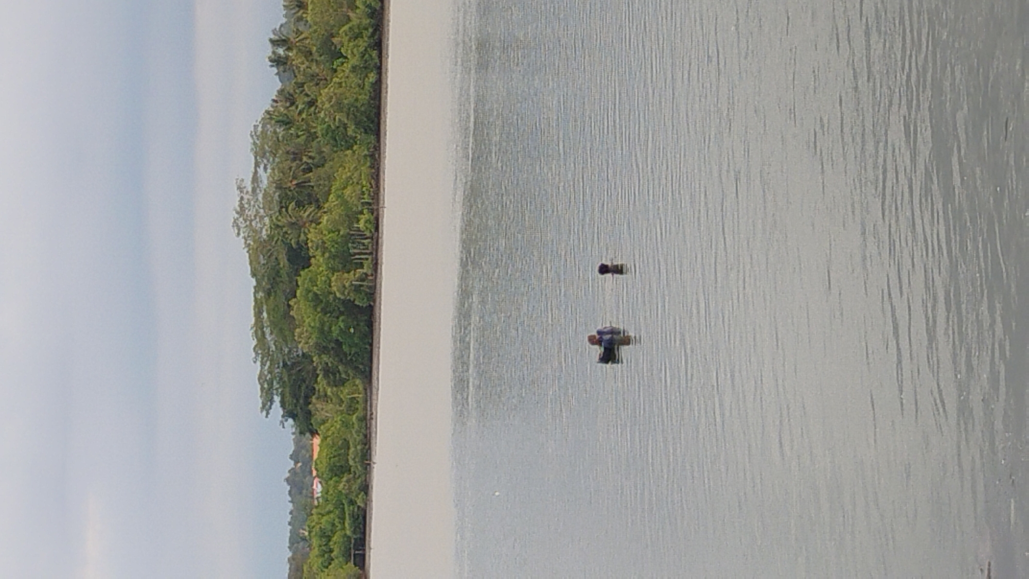 Two individuals are visible in a body of water. One person, wearing a blue shirt, is seated in a black, circular flotation device. The other individual, whose head and shoulders are visible, stands in the water nearby. A tree line with dense green foliage forms the background. The sky is overcast with a light gray color. The location is the Ujong Blang Beach area in Lhokseumawe, Indonesia, known for its coastal environment and mangrove ecosystems.