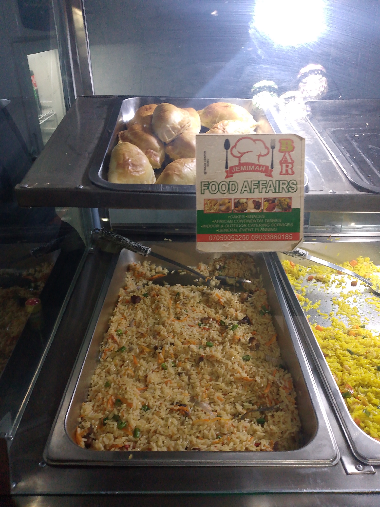 In a brightly lit food display case, a variety of food items are presented in metal trays. The top tray contains a collection of golden-brown pastries, possibly pies or turnovers. Below this, two larger metal trays are filled with rice dishes. The left tray holds a generous portion of fried rice, a beige-colored grain mixed with visible pieces of chopped carrots and green vegetables. To its right, another tray is filled with bright yellow rice, also interspersed with diced vegetables. A white and green sign with a red border is prominently displayed in front of the rice dishes, featuring a chef's hat icon and the text "JEMIMAH FOOD AFFAIRS." The sign also lists services like "CAKES SNACKS," "AFRICAN CONTINENTAL DISHES," and "INDOOR & OUTDOOR CATERING SERVICES." Contact numbers are also visible on the sign. The environment appears to be a food service establishment, with a clean and organized presentation of the food.