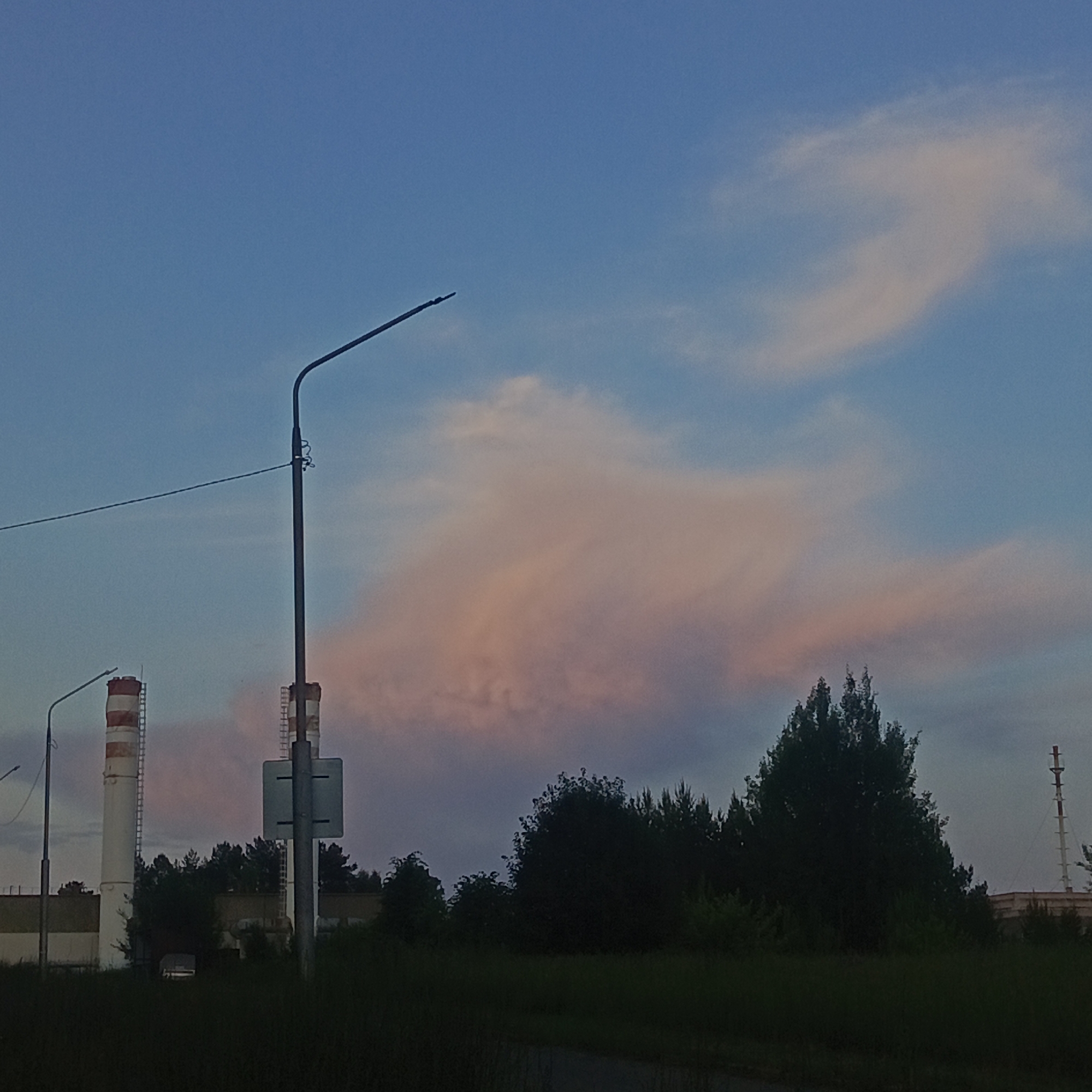 The image depicts a twilight scene in the industrial outskirts of Марковский, Russia. Dominating the sky are wispy clouds tinged with pink and orange hues against a gradient of blue. In the foreground, silhouetted by the fading light, is a dark expanse of grass and foliage. Several elements of industrial infrastructure are visible. To the left, two tall, cylindrical chimneys with red and white stripes stand against the sky, each with a ladder-like structure on its side. A street lamp with a curved arm stands prominently in the center-right of the frame, a wire extending from it horizontally across the upper portion of the image. In the background, a dense line of dark green trees forms a natural barrier. Further to the right, another industrial chimney, taller and thinner than the striped ones, is visible. The overall impression is one of a quiet, almost desolate industrial landscape at dusk.