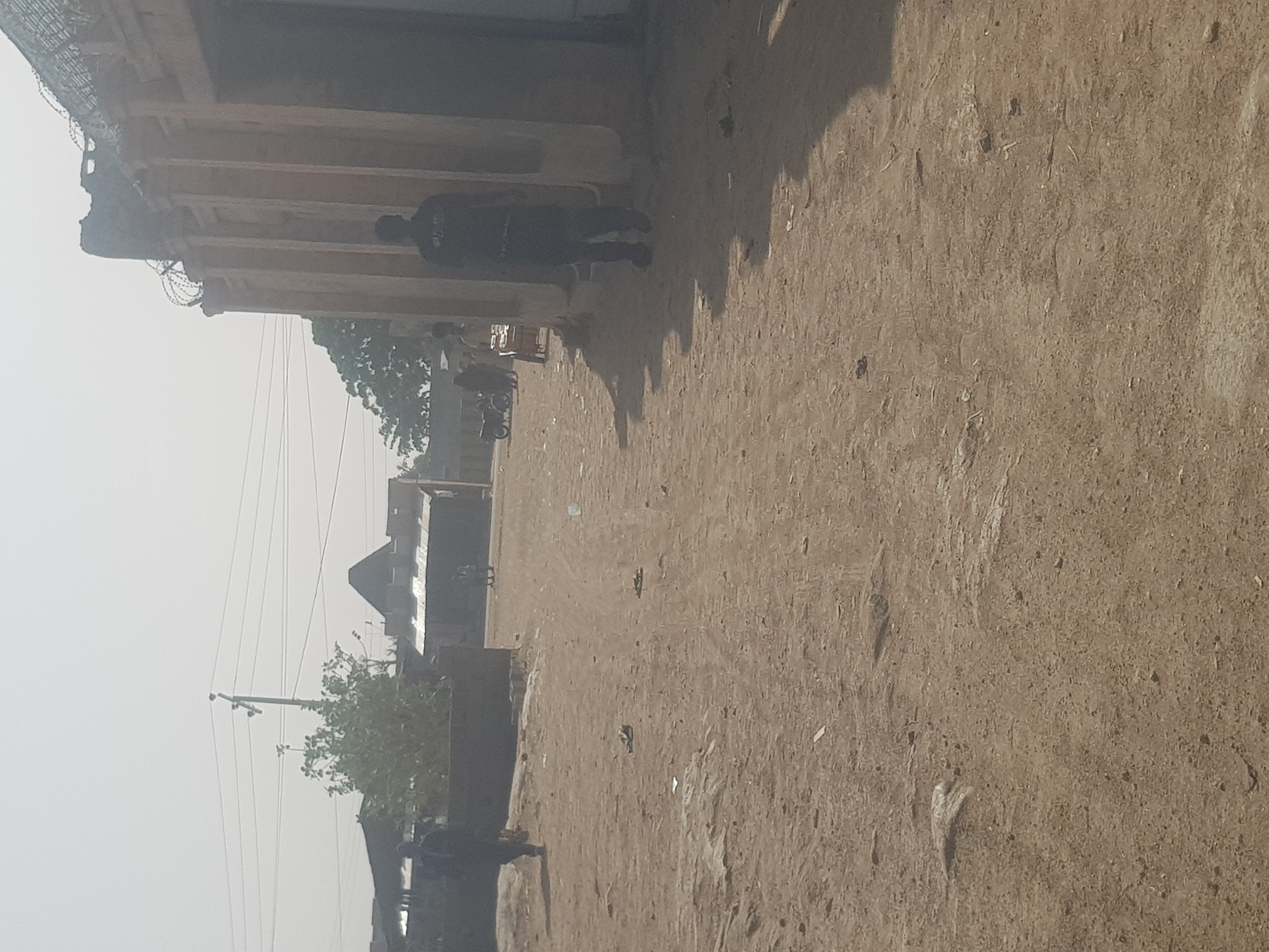 The scene is an outdoor street view in Dutsinma B, Nigeria. The ground is dusty and dirt-covered. A building is located on the left with barbed wire along its upper edge. A person is seen walking along the side of the building. In the distance are other buildings, trees, and electrical poles with wires. The sky is hazy and pale.