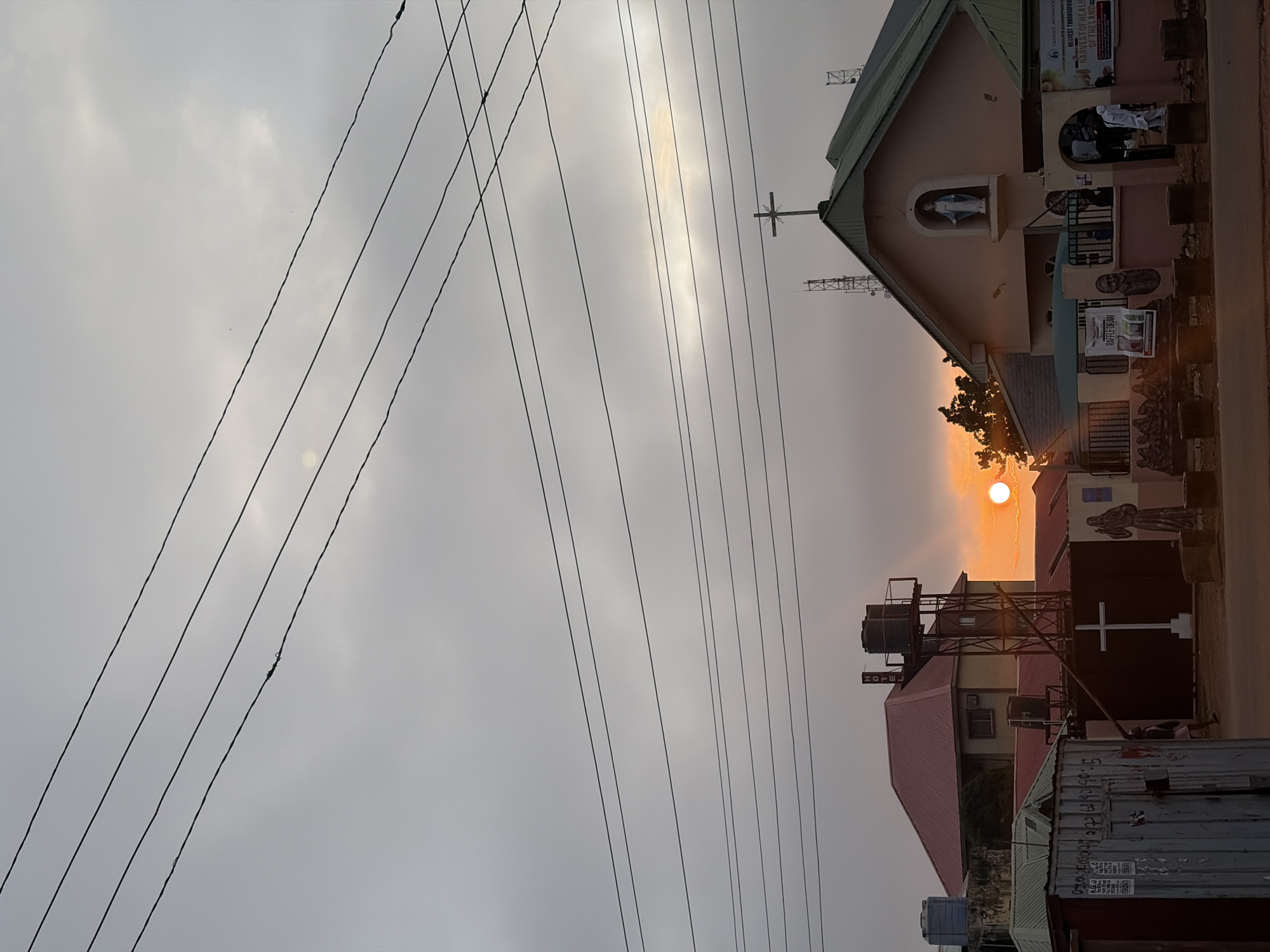 A sunset is occurring in Gura Top, Nigeria, casting a warm orange glow across the sky, partially obscured by a layer of clouds. In the foreground, power lines are strung across the scene, with the sun visible between them and the horizon. Dominating the middle ground is a light-colored church with a green roof, topped by a cross. To the left of the church, a building with a red roof is visible, and further left is a corrugated metal structure with various inscriptions. A large white cross stands in front of a dark fence to the left of the church. A statue of a man, possibly Jesus, is positioned to the right of the cross. Several people are present, though their activities are not clearly discernible due to the distance and lighting. The environment appears to be a street scene with some vegetation and typical urban infrastructure.