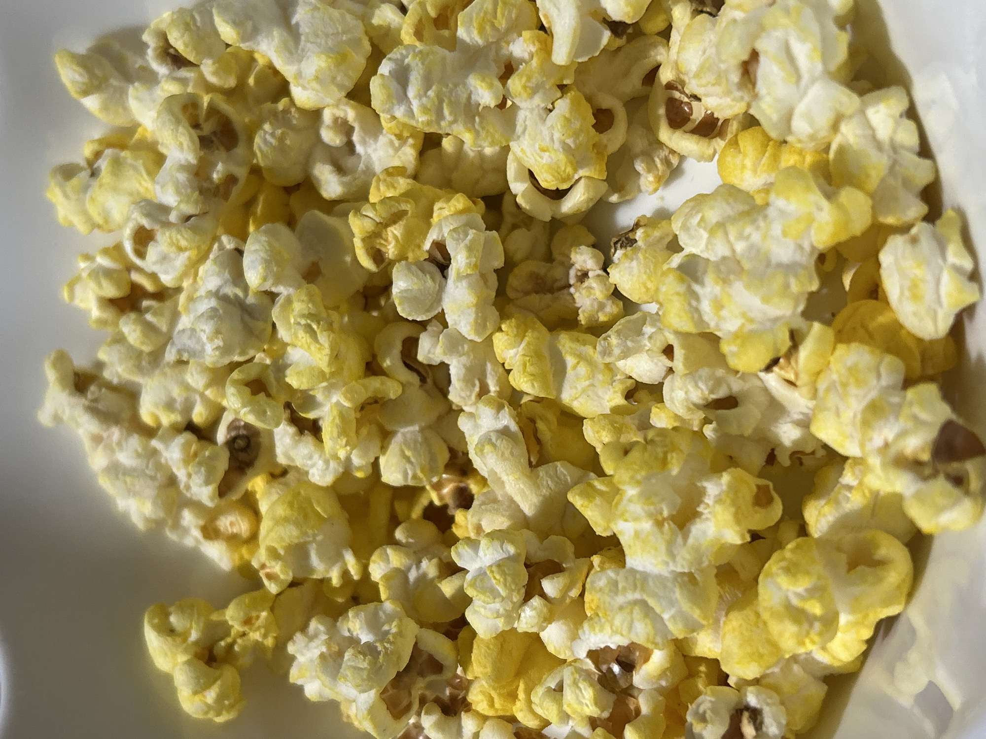 This is a close-up, overhead shot of a white bowl filled with popcorn. The popcorn is a mix of white and bright yellow kernels, with some kernels appearing slightly browned. The lighting suggests an indoor setting, possibly a home. There are no people or any signs of activity, and the background is indistinct, focusing entirely on the popcorn. The image provides no information about the city of Byala Slatina, Bulgaria, or any other geographical location.