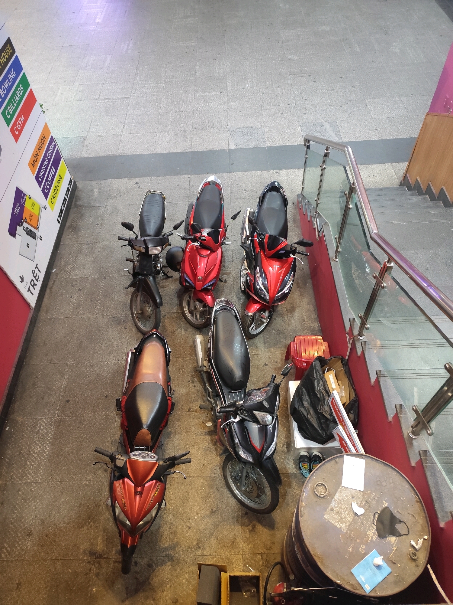 The image, taken from an elevated perspective, depicts a collection of parked motorcycles on a paved surface. To the left, a brightly colored vertical sign displays text in English and Vietnamese, with categories such as "HOUSE," "BOWLING," "C'BILLIARDS," "C'GYM," "MON NGON," "CINESTAR CINEMA," "C'COFFEE," and "C'KIDZONE." This sign, along with the overall aesthetic, suggests the location is within a commercial or entertainment complex in Da Lat, Vietnam.
The motorcycles are predominantly red and black, varying in style from more rugged to sleek, modern designs. One motorcycle, in the foreground, is a vibrant red with black accents. Behind it, a black motorcycle with a prominent silver exhaust pipe is parked. To the right of this, a black motorcycle with red detailing is positioned, facing towards the right side of the frame. Further back, a red motorcycle with black seating is visible, and to its left, a dark-colored motorcycle with a black seat.
Adjacent to the motorcycles on the right side of the image, a metal railing and glass barrier define a set of stairs descending into a lower level. The stairs are painted red, with wooden panels at the bottom. A large, weathered metal barrel, resting on its side, is prominently featured in the lower right corner. On top of the barrel, a black face mask and several pieces of paper are scattered. To the left of the barrel, a black trash bag is placed next to a red object, possibly a piece of luggage or equipment, and a pair of shoes. The ground appears to be paved with concrete tiles. The lighting suggests it is daytime, possibly indoors or under a covered area. No people are visible in the frame.