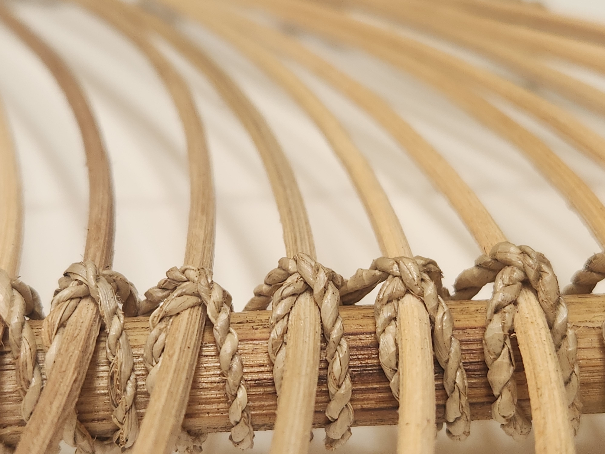 This is a close-up, vertically oriented photograph of woven natural fibers. The dominant feature is a cylindrical, light brown, textured wooden pole around which thick, braided rope is wrapped in a spiraling pattern. The rope is a lighter, beige color with visible individual strands. Intersecting with the pole and rope are several thin, curved, light brown wooden rods or branches that extend outwards and fade into a blurred, off-white background. The lighting is soft and diffused, highlighting the textures of the wood and rope. There are no people, animals, or any discernible elements that would suggest a specific location within the city of Maitland, United States of America. The image focuses solely on the intricate details of the materials and their construction.