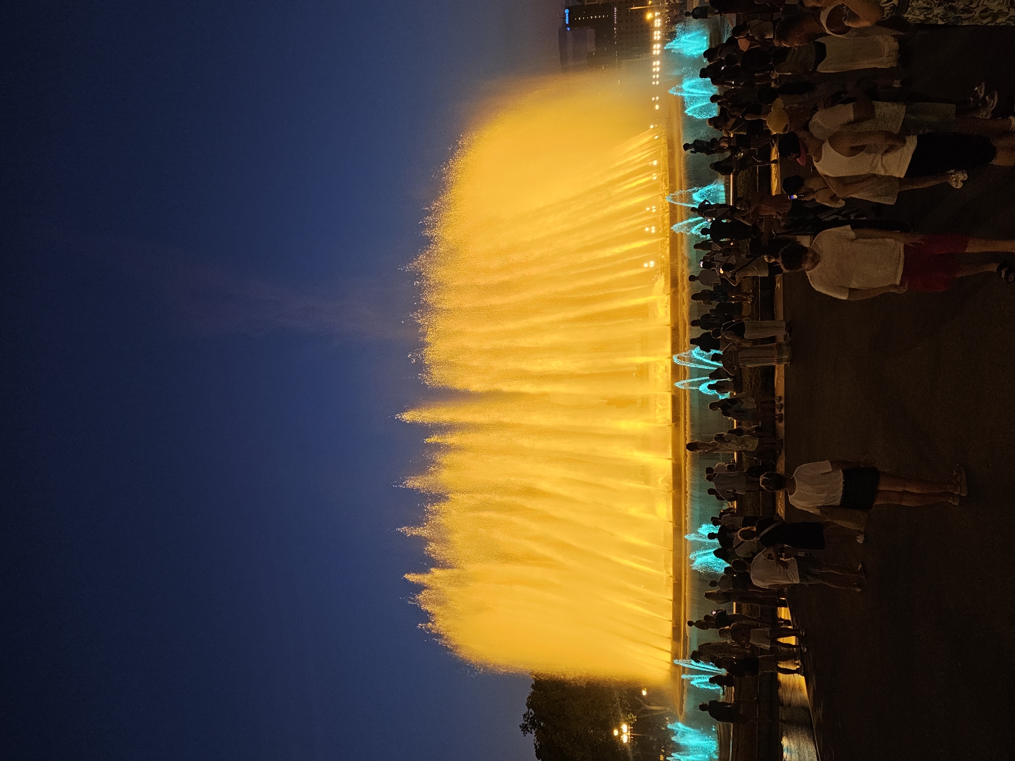 The image captures a nighttime view of the Magic Fountain of Montjuïc in Barcelona, Spain. A large fountain, illuminated with bright yellow and orange lights, dominates the center of the frame, with multiple streams of water shooting upwards. Smaller, turquoise-colored fountains are visible in the foreground and to the sides. A crowd of people is gathered in front of the fountain, observing the spectacle. They are dressed in casual summer clothing. The sky above is a deep, dark blue, indicating it is after sunset. Trees and some distant buildings are visible on the left and right edges of the scene, respectively. The ground in the foreground appears to be wet, reflecting the fountain's lights.