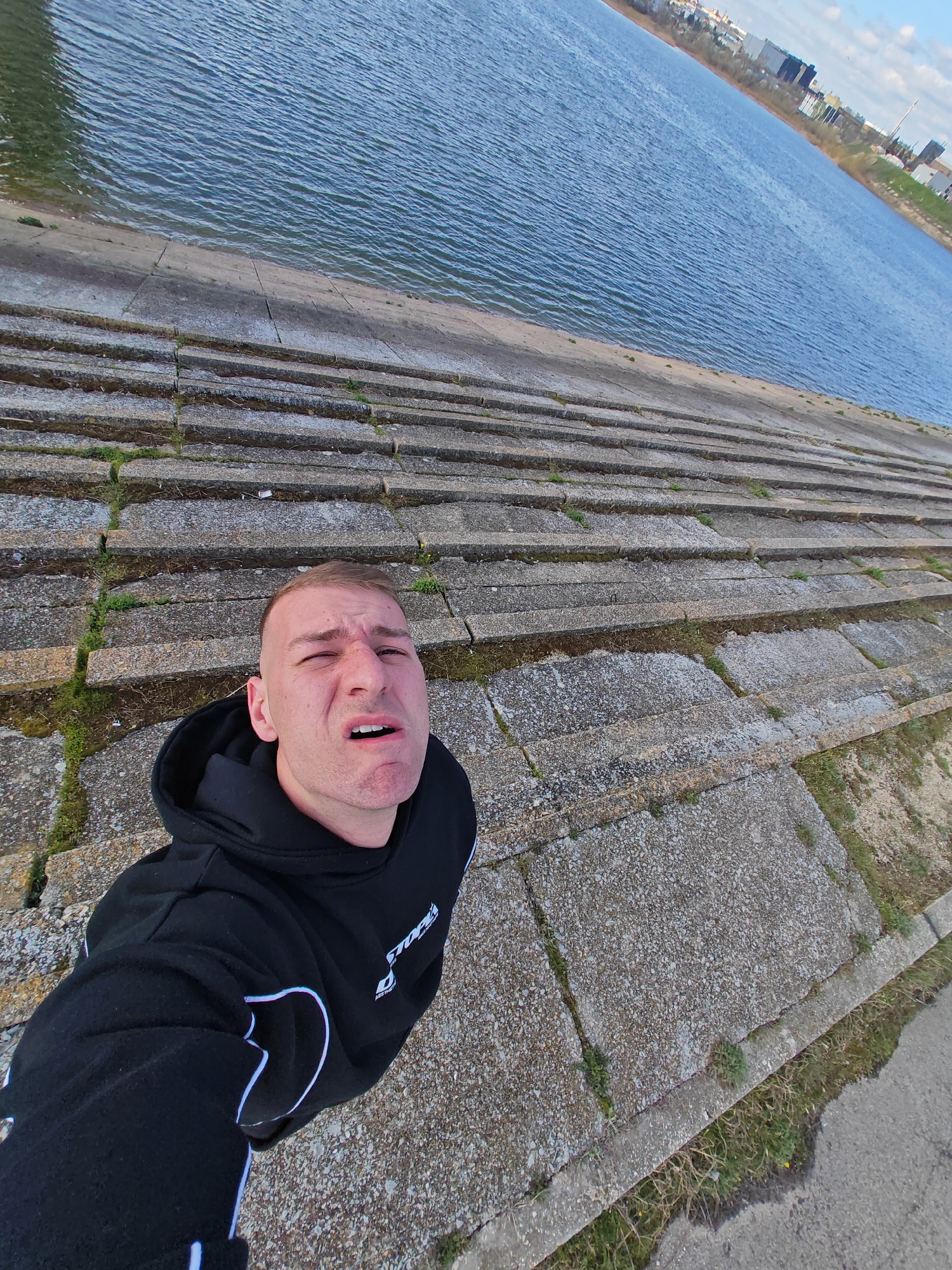 A man is taking a selfie on a stepped concrete embankment next to a body of water. The water is blue with small ripples. In the distance, there are buildings and trees. The man is wearing a black hoodie with white accents. The concrete steps have some green moss and vegetation growing between the cracks. The location could be in the city of Shumen, Bulgaria.